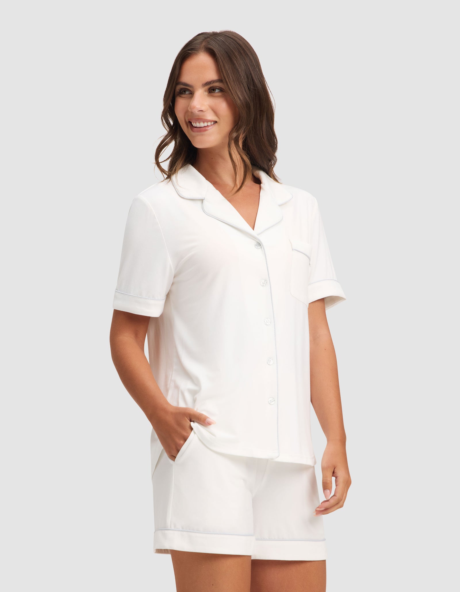 A woman smiles with her hands in her pockets, modeling the Cozy Earth Women's Bamboo Stretch-Knit Short Sleeve Pajama Set—a white button-up top and shorts—against a plain light gray background. 