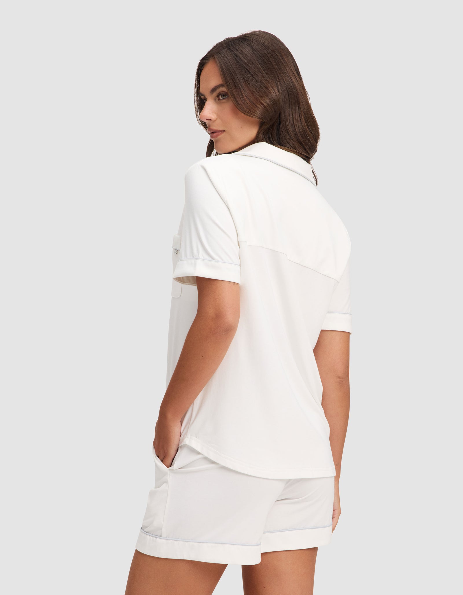 A woman with long brown hair stands sideways, looking over her shoulder, wearing the Cozy Earth Women's Bamboo Stretch-Knit Short Sleeve Pajama Set—loose-fitting white shirt and matching shorts, hands in pockets, set against a plain gray background. 