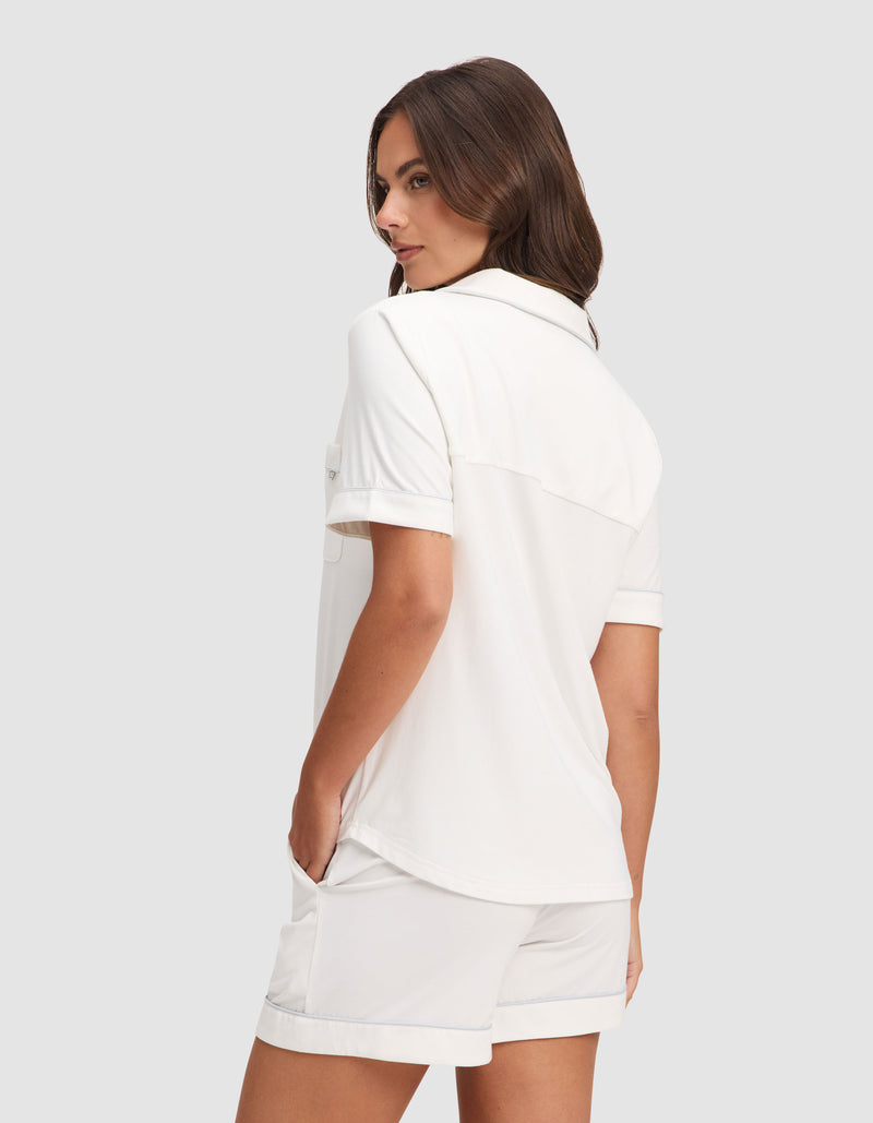 A woman with long brown hair stands sideways, looking over her shoulder, wearing the Cozy Earth Women's Bamboo Stretch-Knit Short Sleeve Pajama Set—loose-fitting white shirt and matching shorts, hands in pockets, set against a plain gray background.