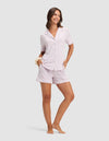 A woman smiles barefoot, wearing Cozy Earth’s Women's Bamboo Stretch-Knit Short Sleeve Pajama Set in light purple—featuring a button-up shirt and matching shorts, with a scrunchie on her wrist and one hand on her hip against a plain background. |Color:Lilac