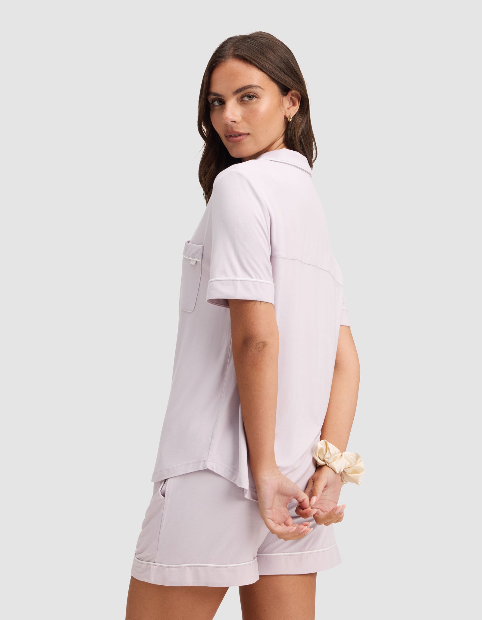 A woman models the Cozy Earth Women's Bamboo Stretch-Knit Short Sleeve Pajama Set in light lavender, standing with her back slightly turned, hands clasped behind her, and a beige scrunchie on her wrist. 