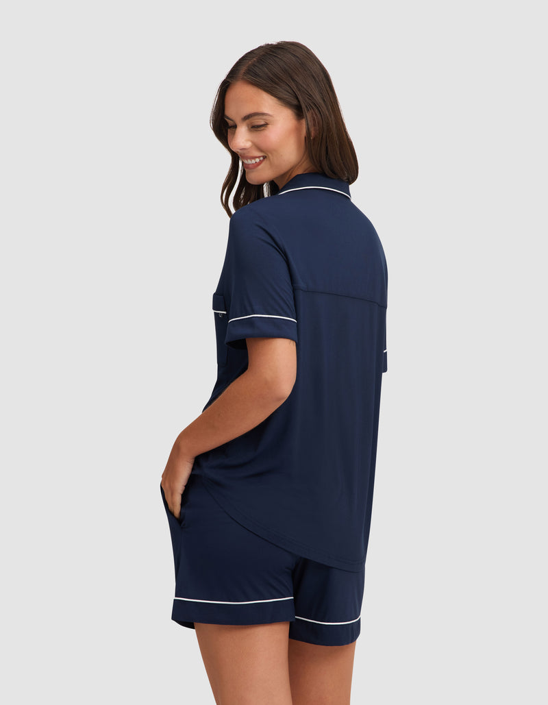 A woman with long brown hair models the Cozy Earth Women's Bamboo Stretch-Knit Short Sleeve Pajama Set in navy blue with white piping, featuring a button-up top and shorts. She smiles, glancing over her shoulder against a light gray background.