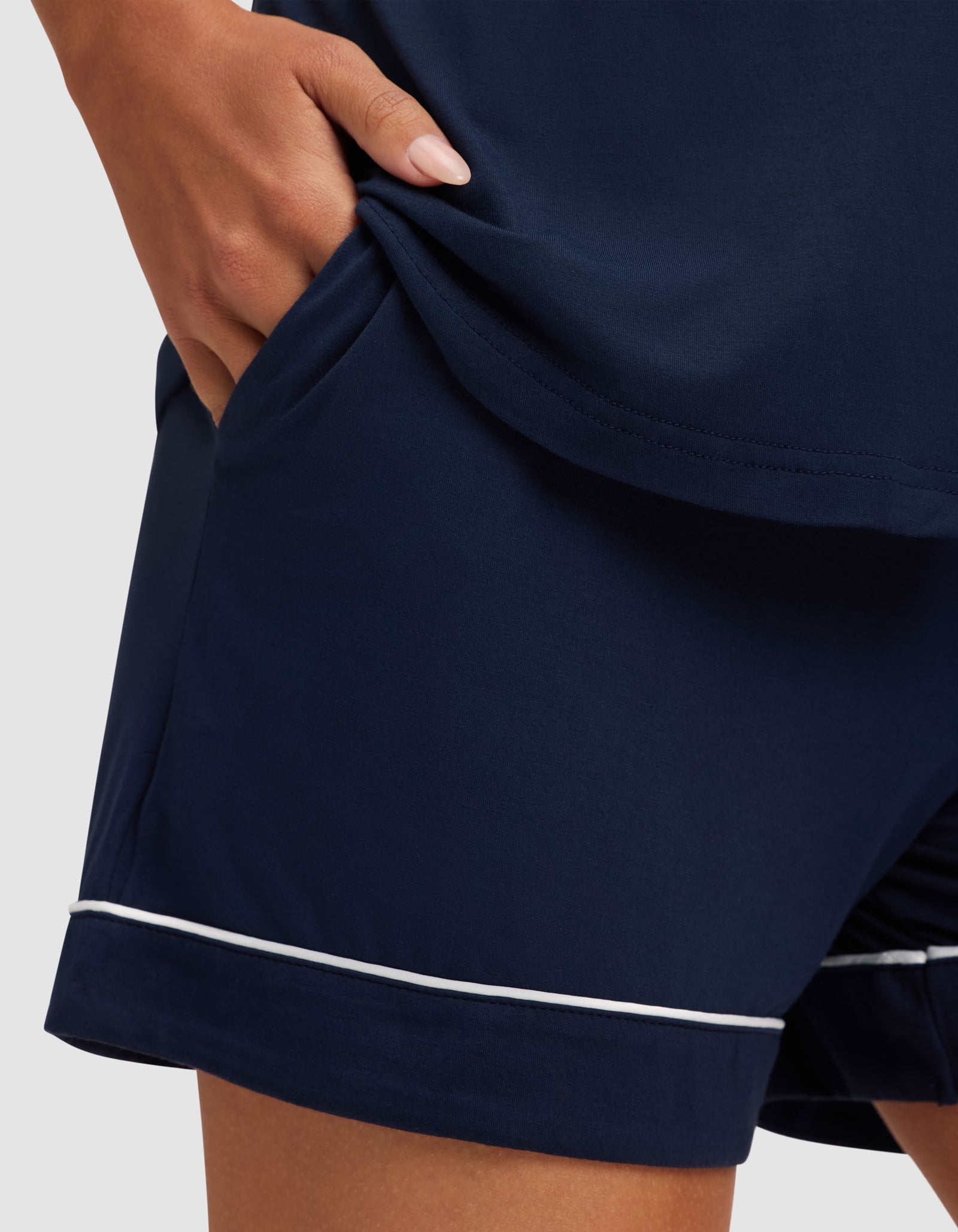 A woman models the Cozy Earth Women's Bamboo Stretch-Knit Short Sleeve Pajama Set, featuring navy blue shorts with white piping and a hand in the pocket, posed against a plain, light-colored background. 