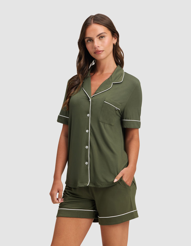 A woman wears the Cozy Earth Women's Bamboo Stretch-Knit Short Sleeve Pajama Set in dark green with white piping and buttons, standing against a plain light gray background.