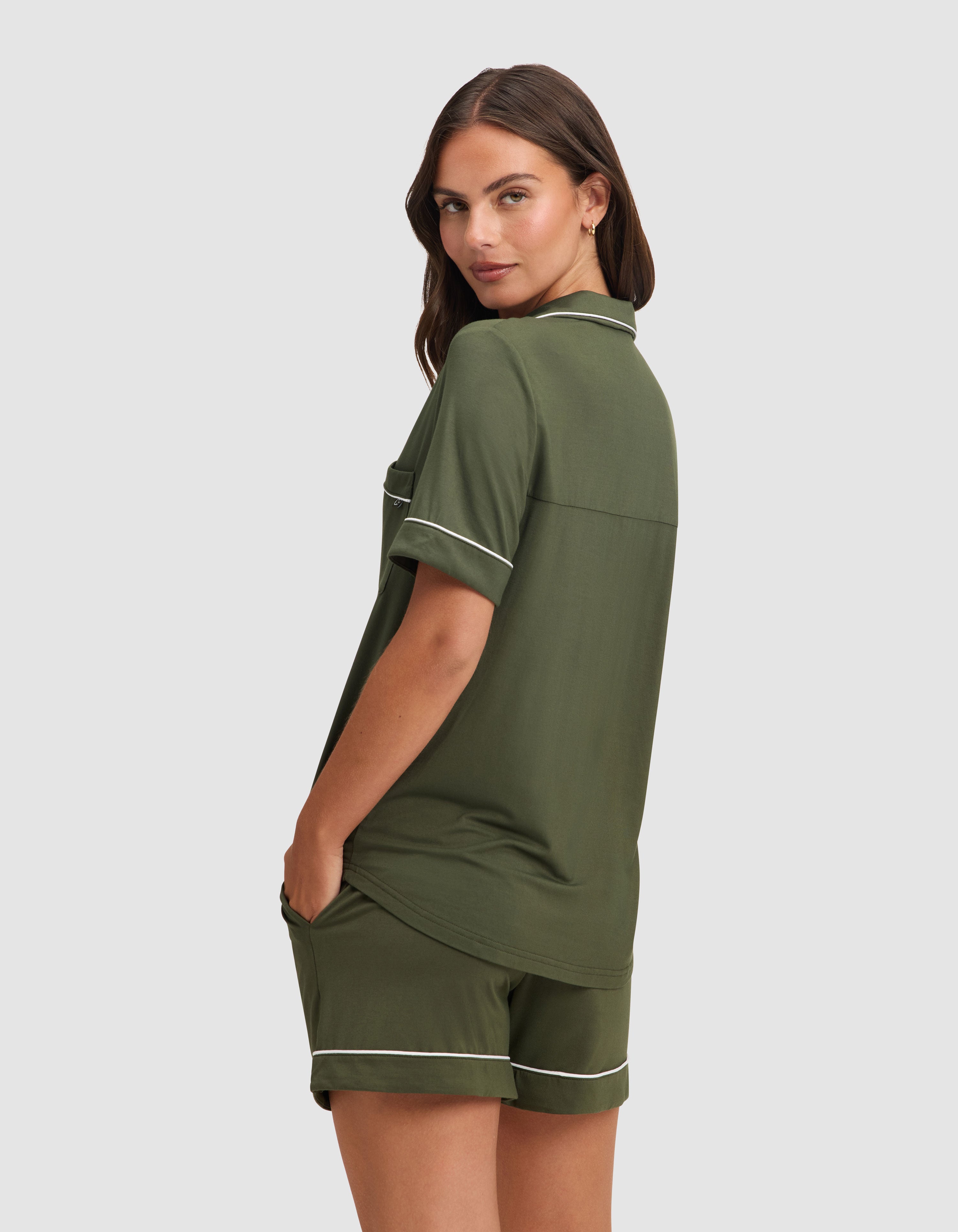 A woman models the Cozy Earth Women's Bamboo Stretch-Knit Short Sleeve Pajama Set in olive green with white piping, standing sideways with hands in her shorts' pockets, against a plain light gray background. |Color:Olive