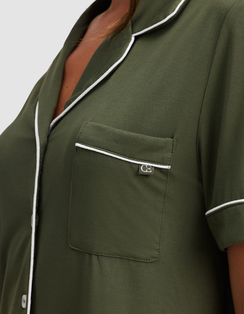 Close-up of a person wearing Cozy Earth's Women's Bamboo Stretch-Knit Short Sleeve Pajama Set in olive green, featuring white piping on the collar, pocket, and sleeves, a small chest pocket with silver tag, and white front buttons.