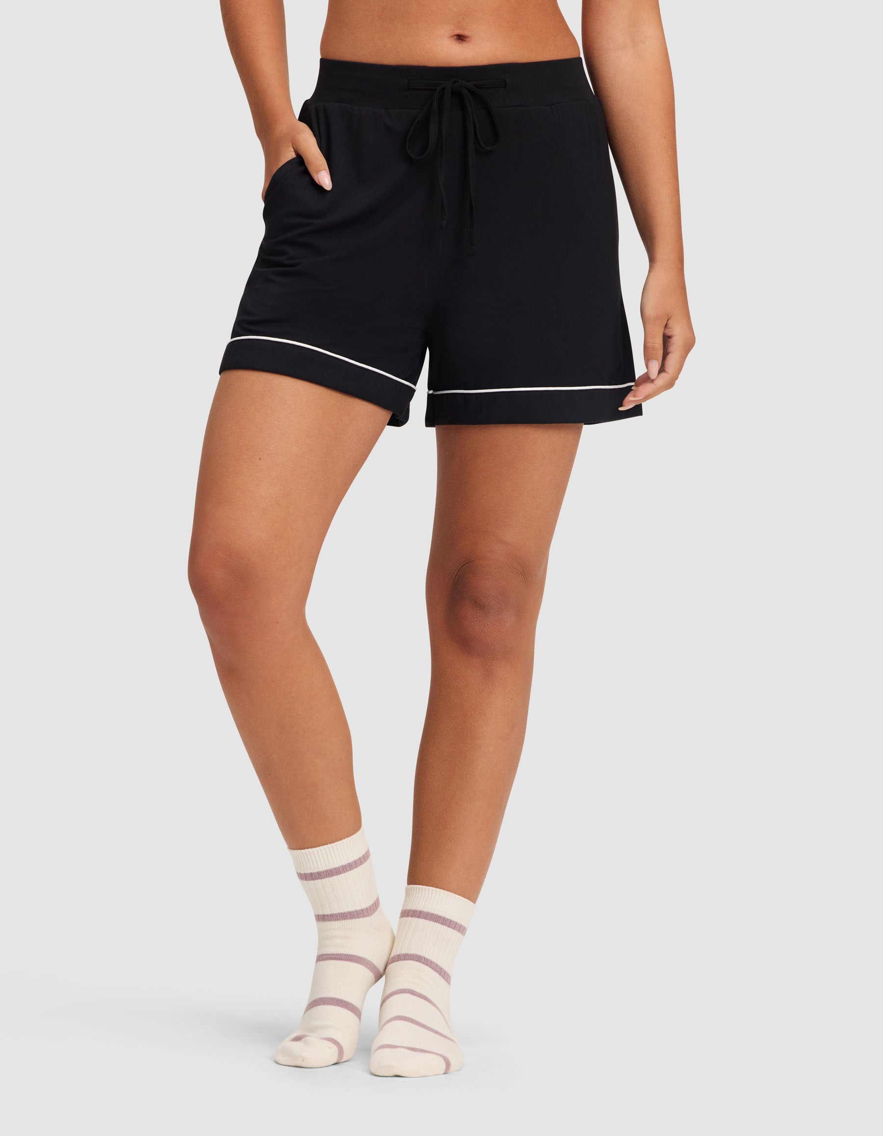 A person wears HIDE Women's Bamboo Stretch-Knit Pajama Shorts in black with white trim and light-colored striped socks, standing against a plain light gray background. 