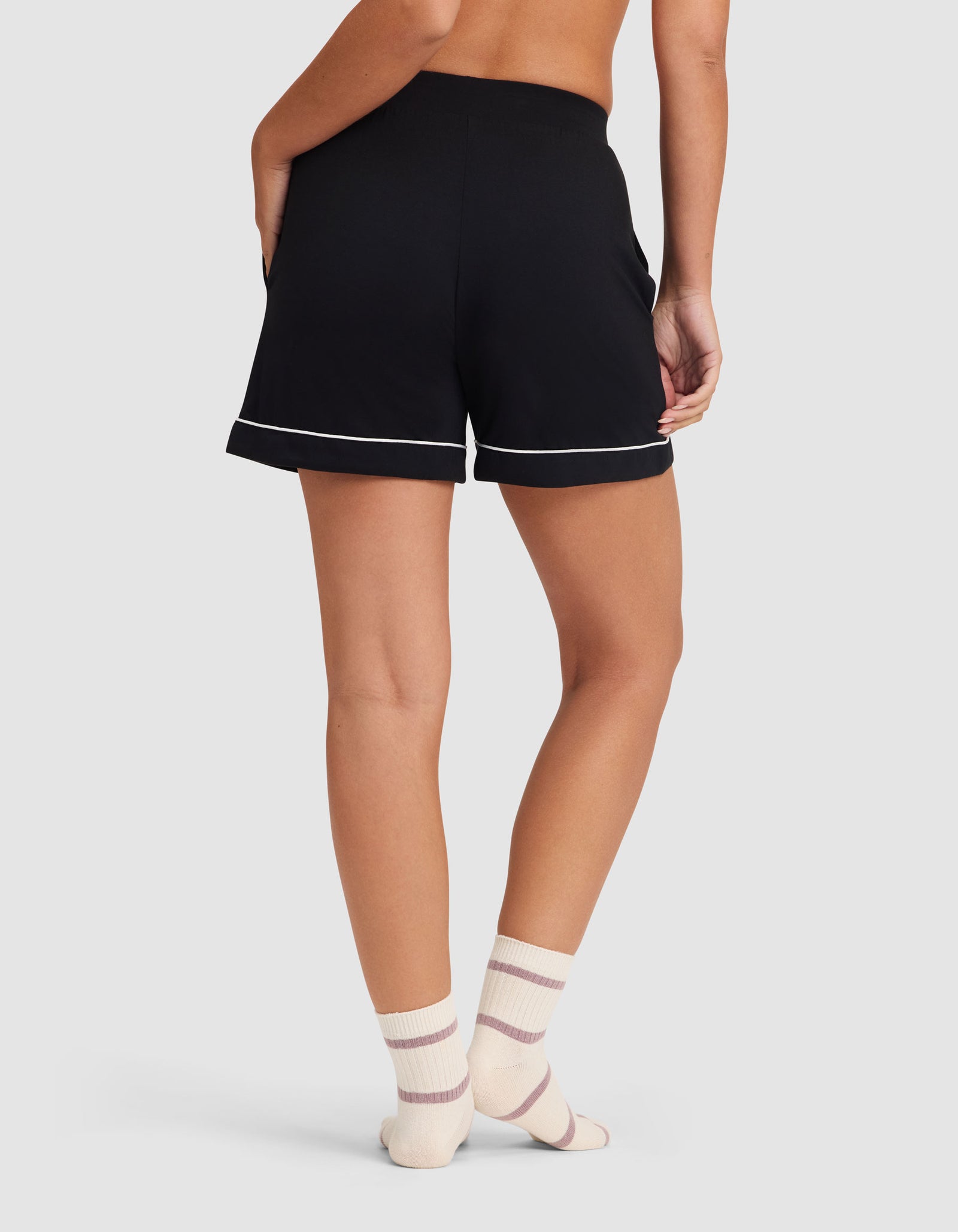 A person faces away, wearing the HIDE Women's Bamboo Stretch-Knit Pajama Short in black with white trim and cream socks with pink stripes, set against a plain light background. 