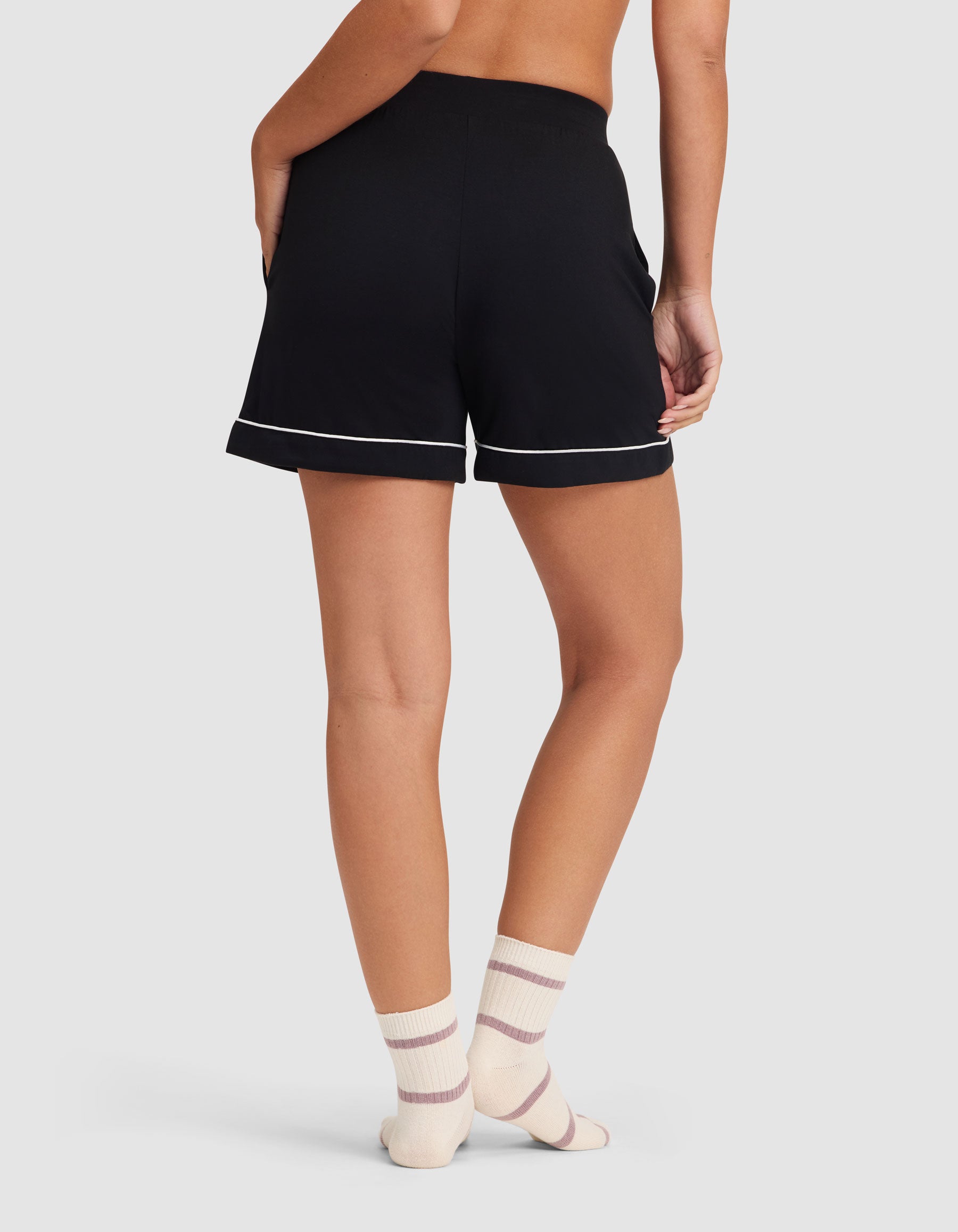 A person faces away, wearing the HIDE Women's Bamboo Stretch-Knit Pajama Short in black with white trim and cream socks with pink stripes, set against a plain light background. 