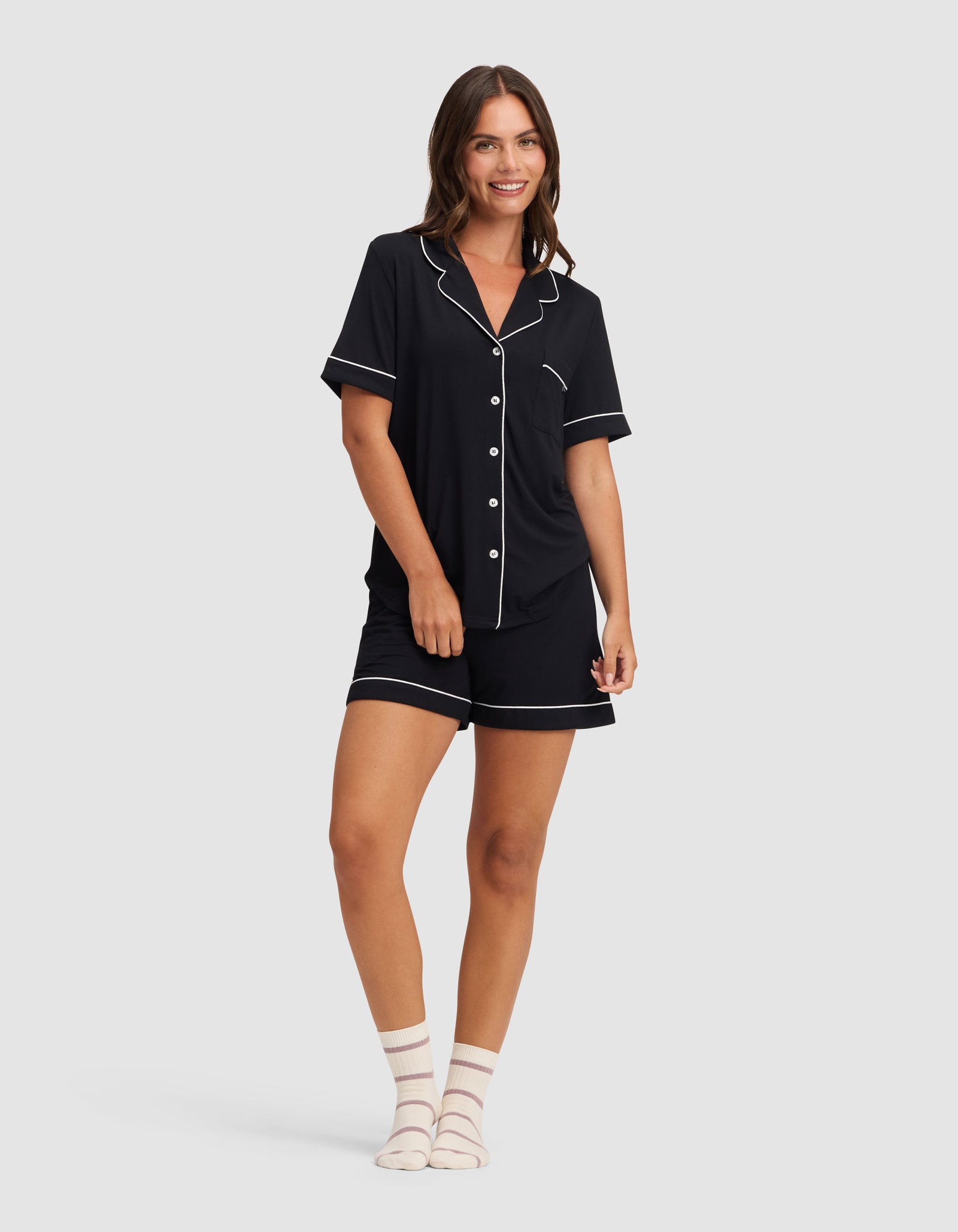 A woman smiles, wearing HIDE's Women's Bamboo Stretch-Knit Pajama Short set in black with white piping—a short-sleeve button-up shirt and shorts—paired with white ankle socks featuring light pink stripes. 