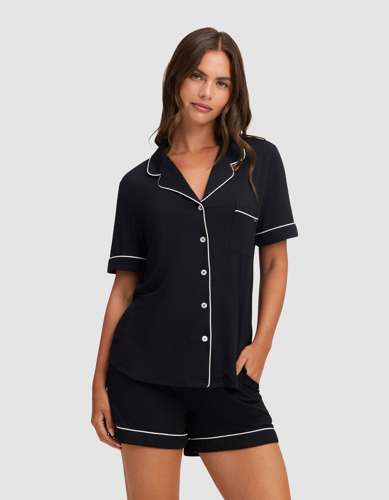 Against a light gray background, a woman wears HIDE's Women's Bamboo Stretch-Knit Pajama Short set in black with white piping and buttons, one hand in her shorts pocket and a relaxed, neutral expression.