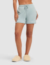 A person stands against a plain white background wearing HIDE Women's Bamboo Stretch-Knit Pajama Shorts in light blue with white trim and beige fuzzy slippers.  |Color:Brook