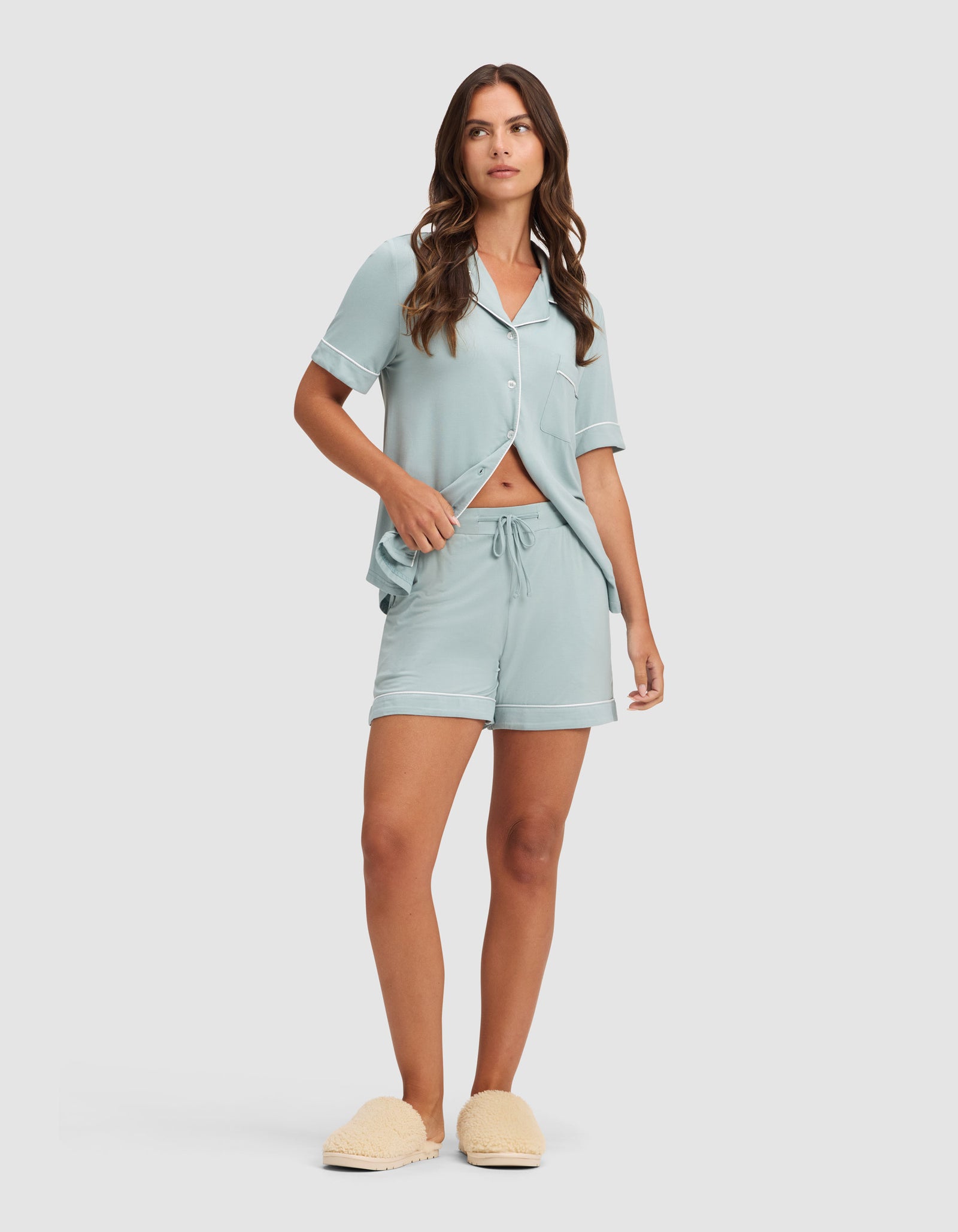 A woman with long brown hair wears HIDE's Women's Bamboo Stretch-Knit Pajama Short set in light blue, featuring a short-sleeve button-up top and matching drawstring shorts, paired with beige slippers against a plain light background. 