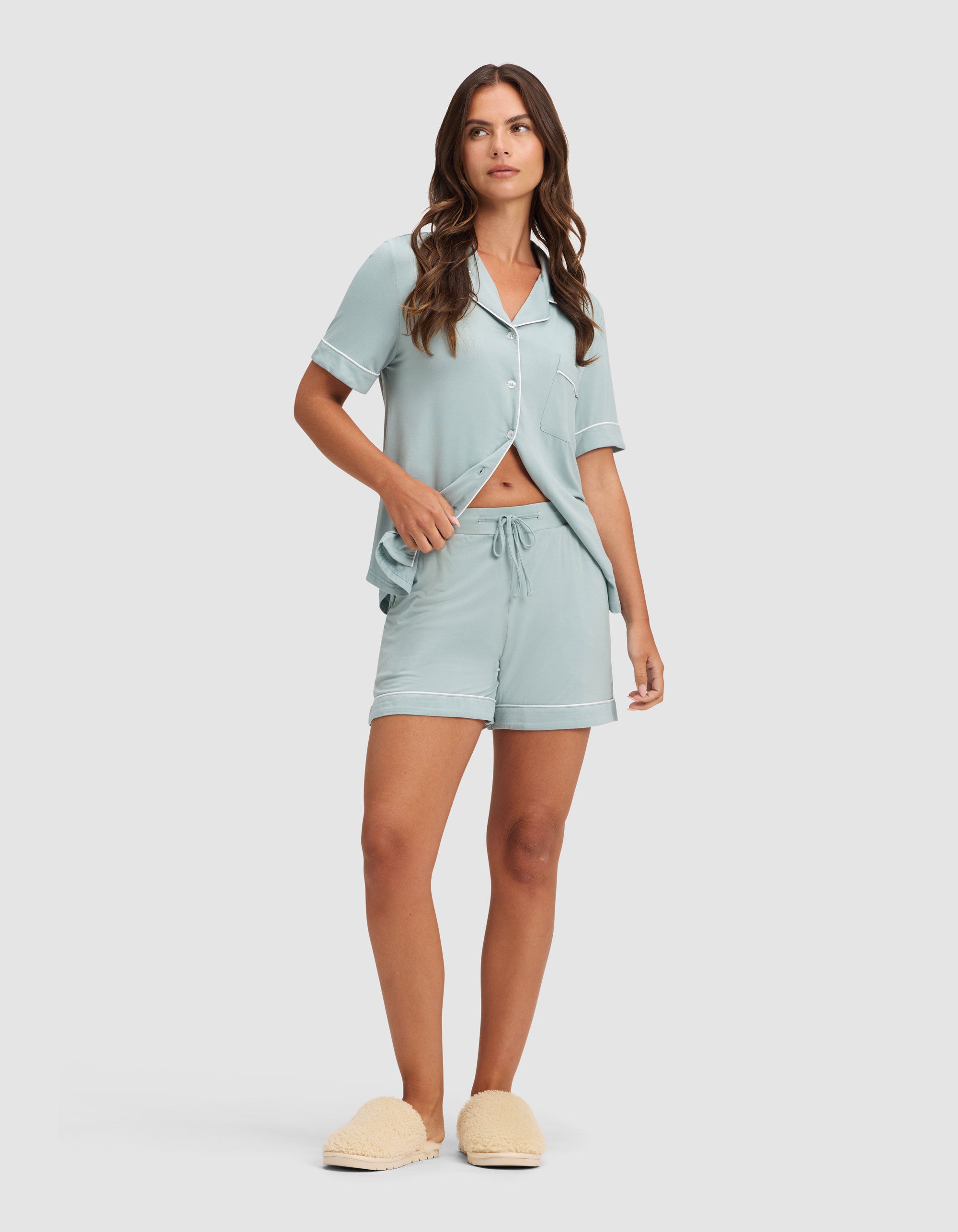 A woman with long brown hair wears HIDE's Women's Bamboo Stretch-Knit Pajama Short set in light blue, featuring a short-sleeve button-up top and matching drawstring shorts, paired with beige slippers against a plain light background. 