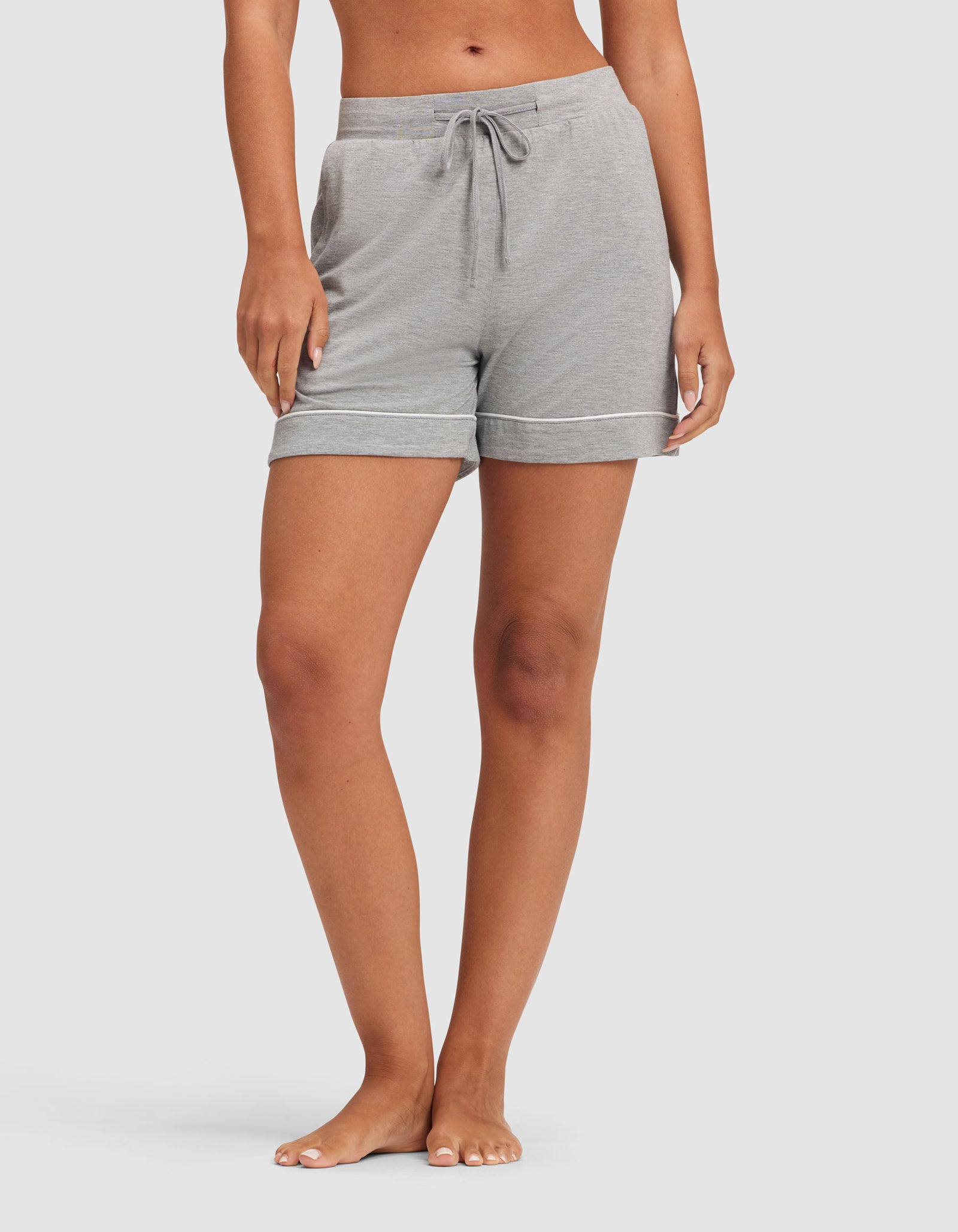 A model, shown from shoulders down, stands barefoot against a white background wearing HIDE's Women's Bamboo Stretch-Knit Pajama Short in light gray with pockets and a drawstring waist. 