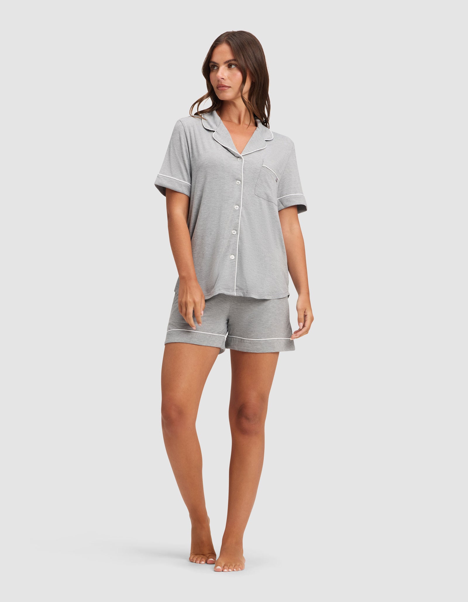 A woman stands barefoot against a white background wearing HIDE's Women's Bamboo Stretch-Knit Pajama Short set in light grey, featuring a button-up, collared short-sleeve top and matching shorts. 