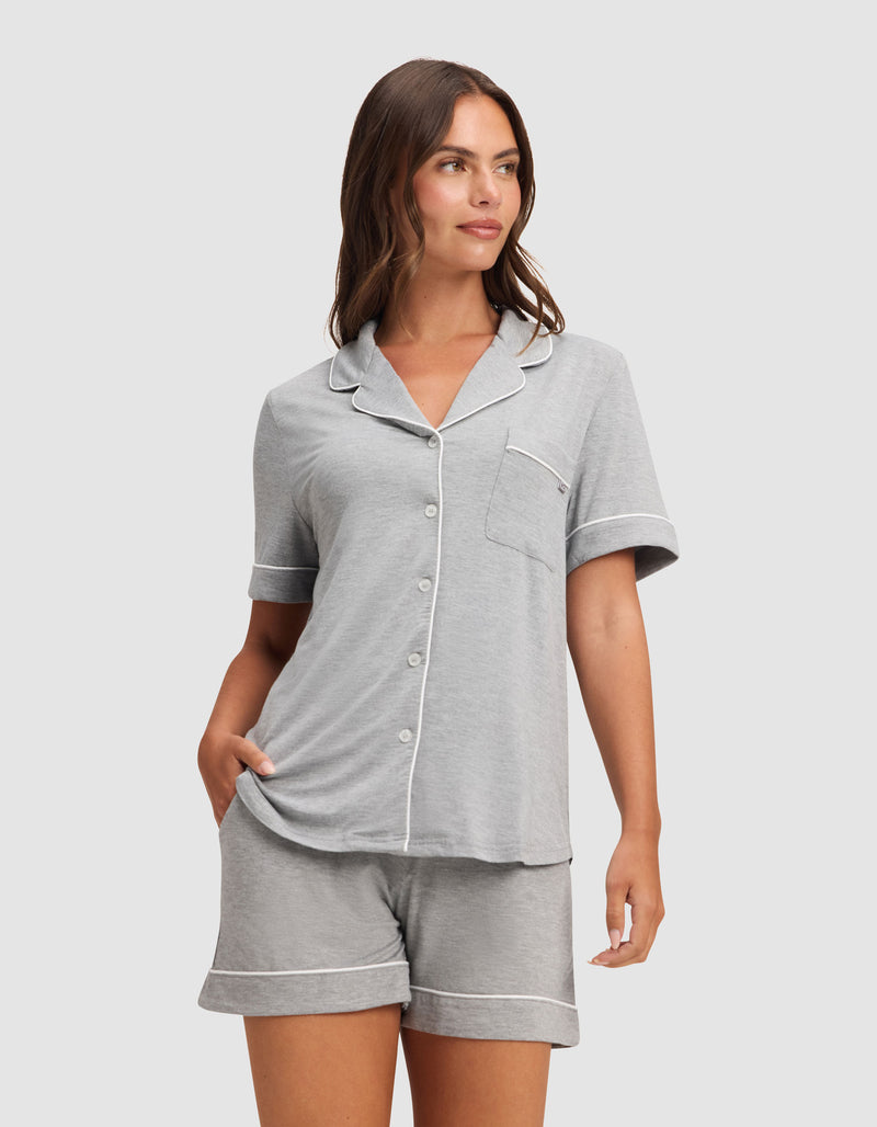 A woman models HIDE's Women's Bamboo Stretch-Knit Pajama Short, featuring a light gray short-sleeve button-up top with white piping and matching shorts, standing with one hand in her pocket against a plain background.