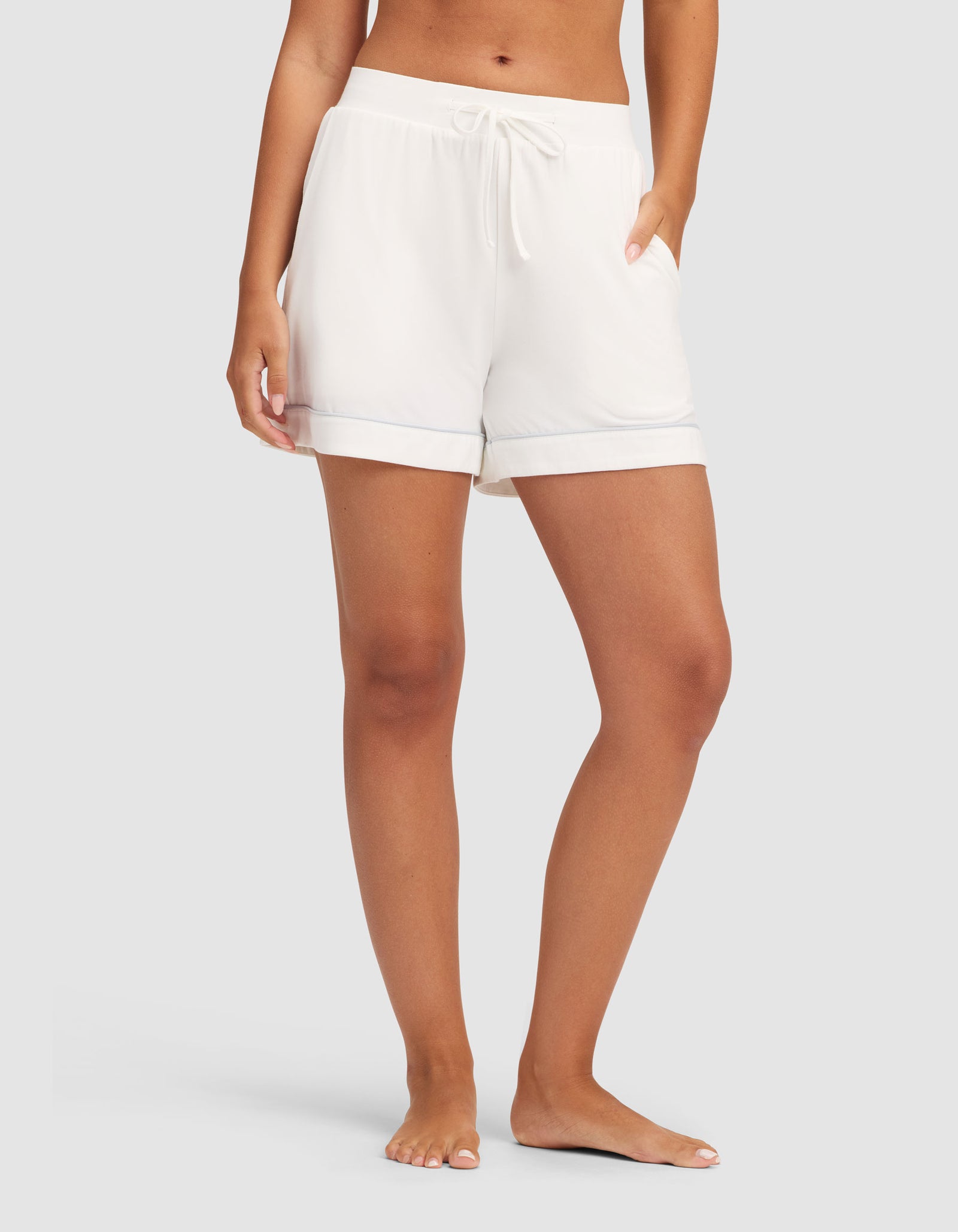 A person stands barefoot against a light gray background, wearing HIDE Women's Bamboo Stretch-Knit Pajama Shorts with rolled cuffs and side pockets; the upper body is mostly out of frame. 