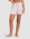 A person stands barefoot against a plain light gray background, wearing HIDE Women's Bamboo Stretch-Knit Pajama Shorts in light purple. The mid-thigh shorts have a relaxed fit and drawstring waist; only the lower body is visible. |Color:Lilac