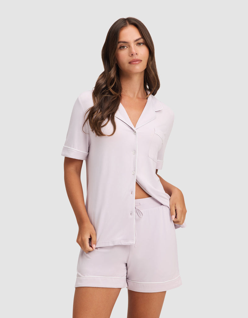 A woman with long brown hair wears the HIDE Women's Bamboo Stretch-Knit Pajama Short set with a button-up top and matching shorts in light lavender, standing against a neutral background.