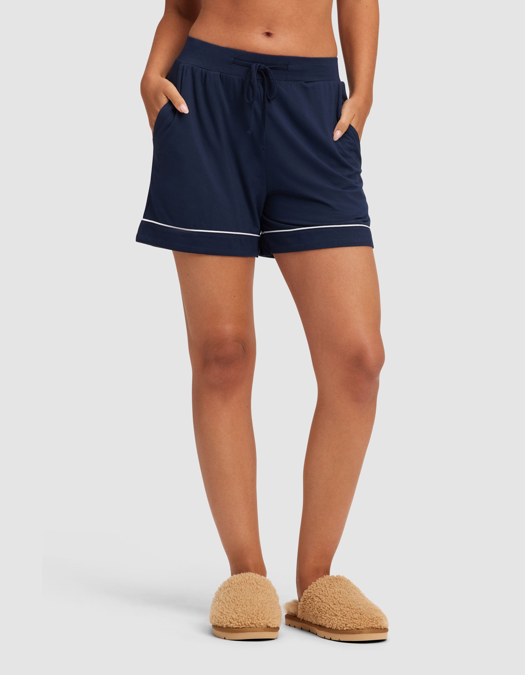 A woman wears HIDE Women's Bamboo Stretch-Knit Pajama Shorts in navy with white piping, hands in pockets, and tan fuzzy slippers, standing against a plain light background.
