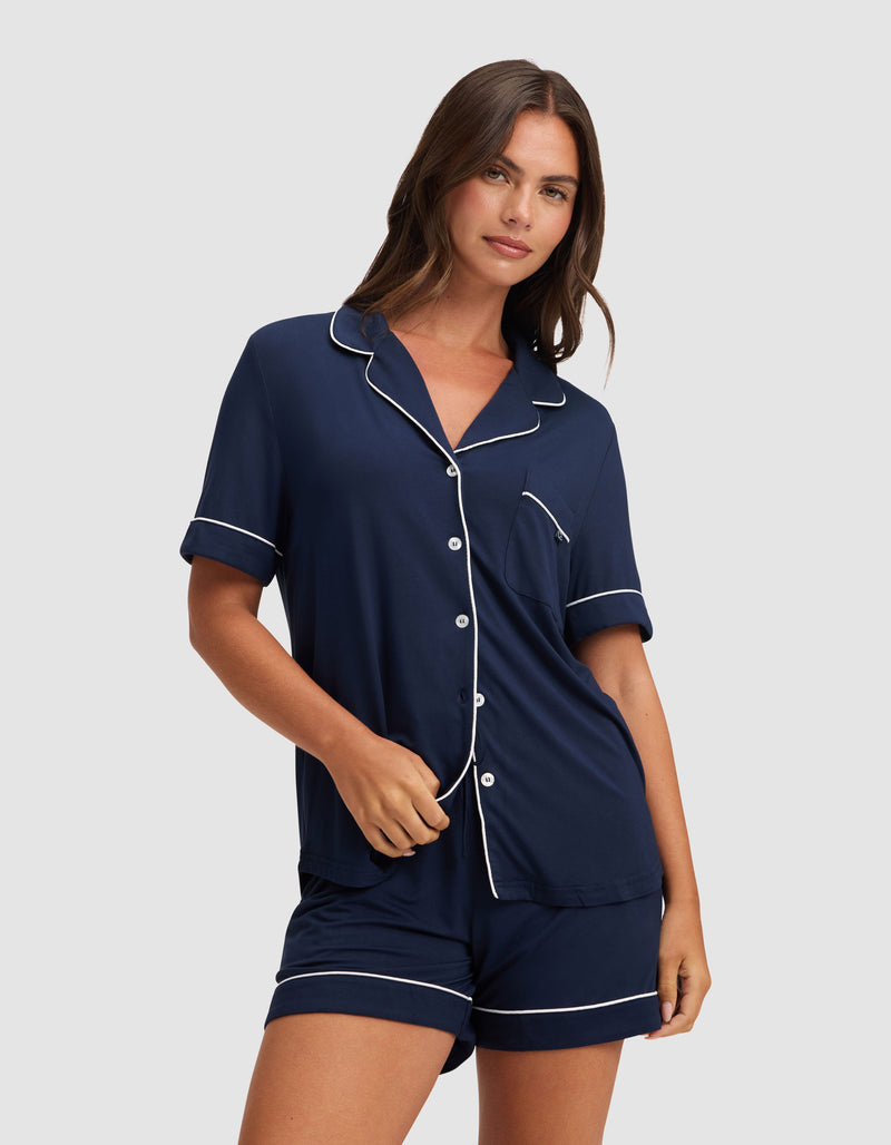 A woman models the Cozy Earth Women's Bamboo Stretch-Knit Pajama Short set in navy blue with white piping, featuring a short-sleeve button-up top and matching shorts, against a plain light gray background.