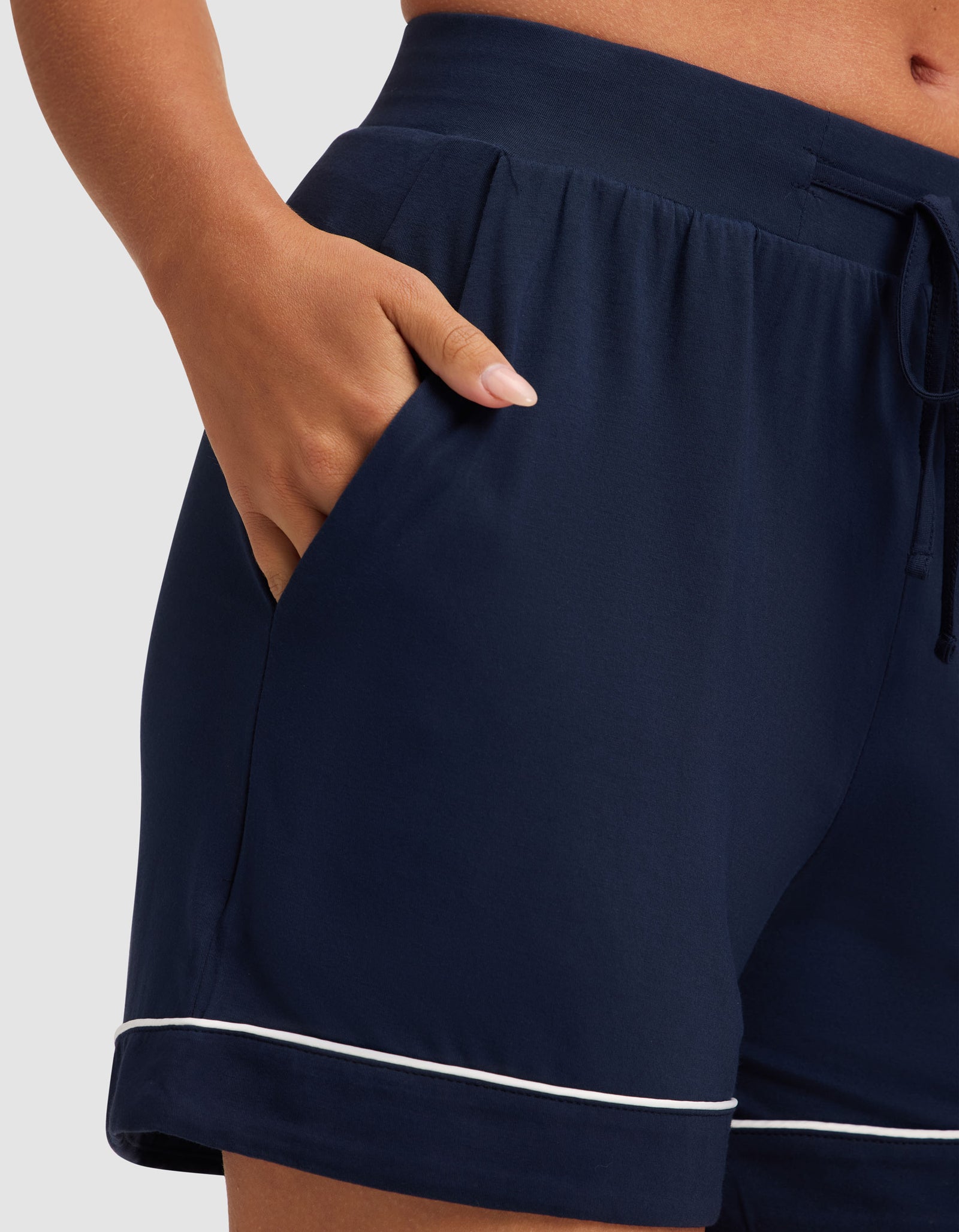 A woman wears HIDE's Women's Bamboo Stretch-Knit Pajama Short in navy with white piping near the hem, shown waist to mid-thigh, one hand in her pocket. 