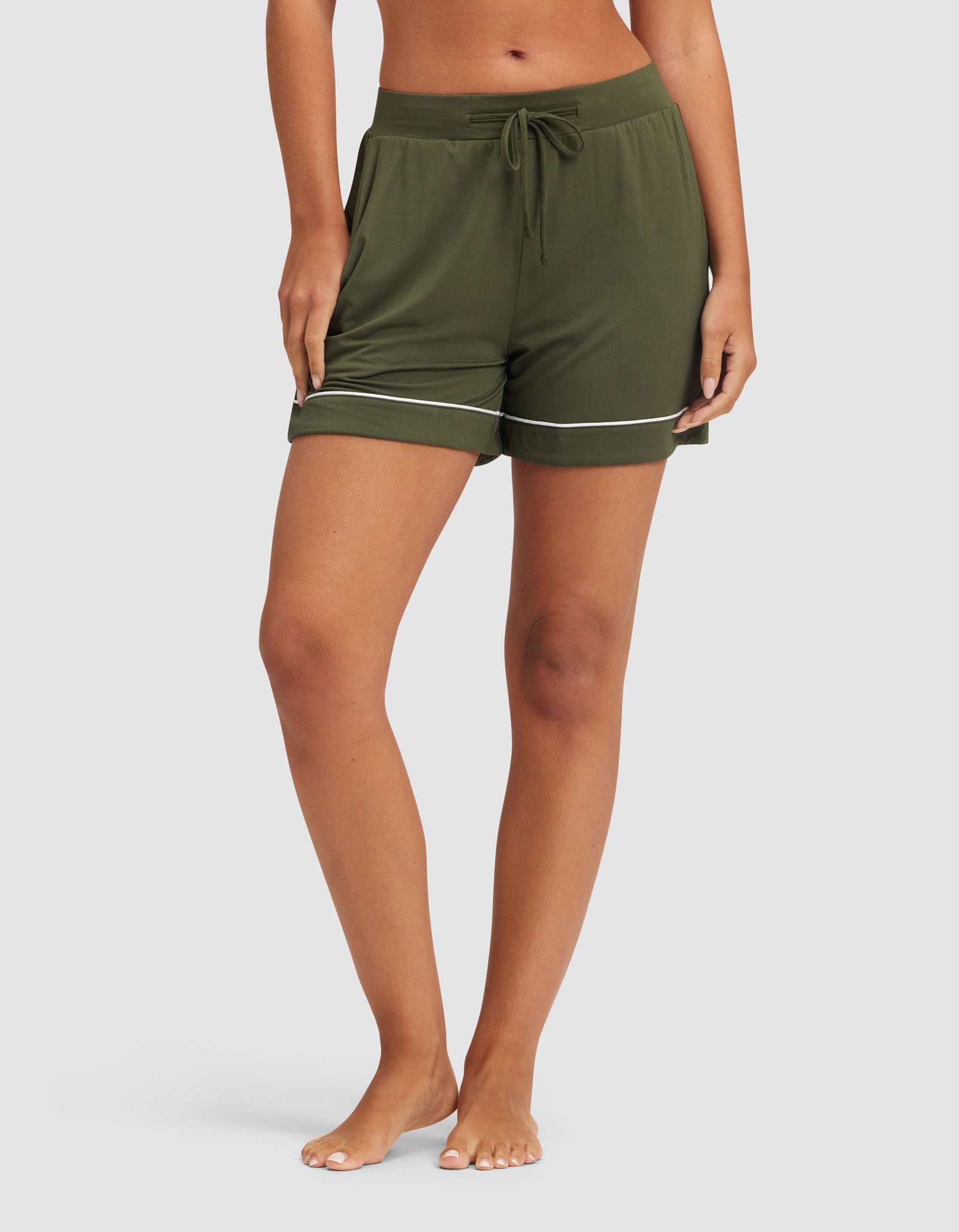 A woman stands barefoot on a light background, hands in the pockets of HIDE Women's Bamboo Stretch-Knit Pajama Shorts in olive green with white trim. 