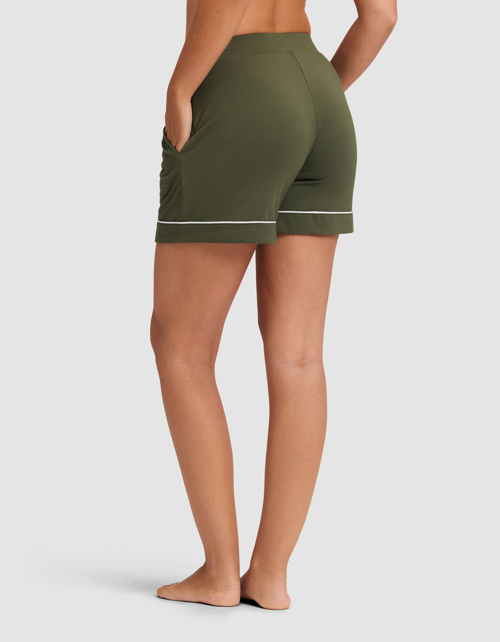A woman models HIDE's Women's Bamboo Stretch-Knit Pajama Short in olive green with white piping, standing barefoot on a white background, viewed from the back with one hand in a pocket. 