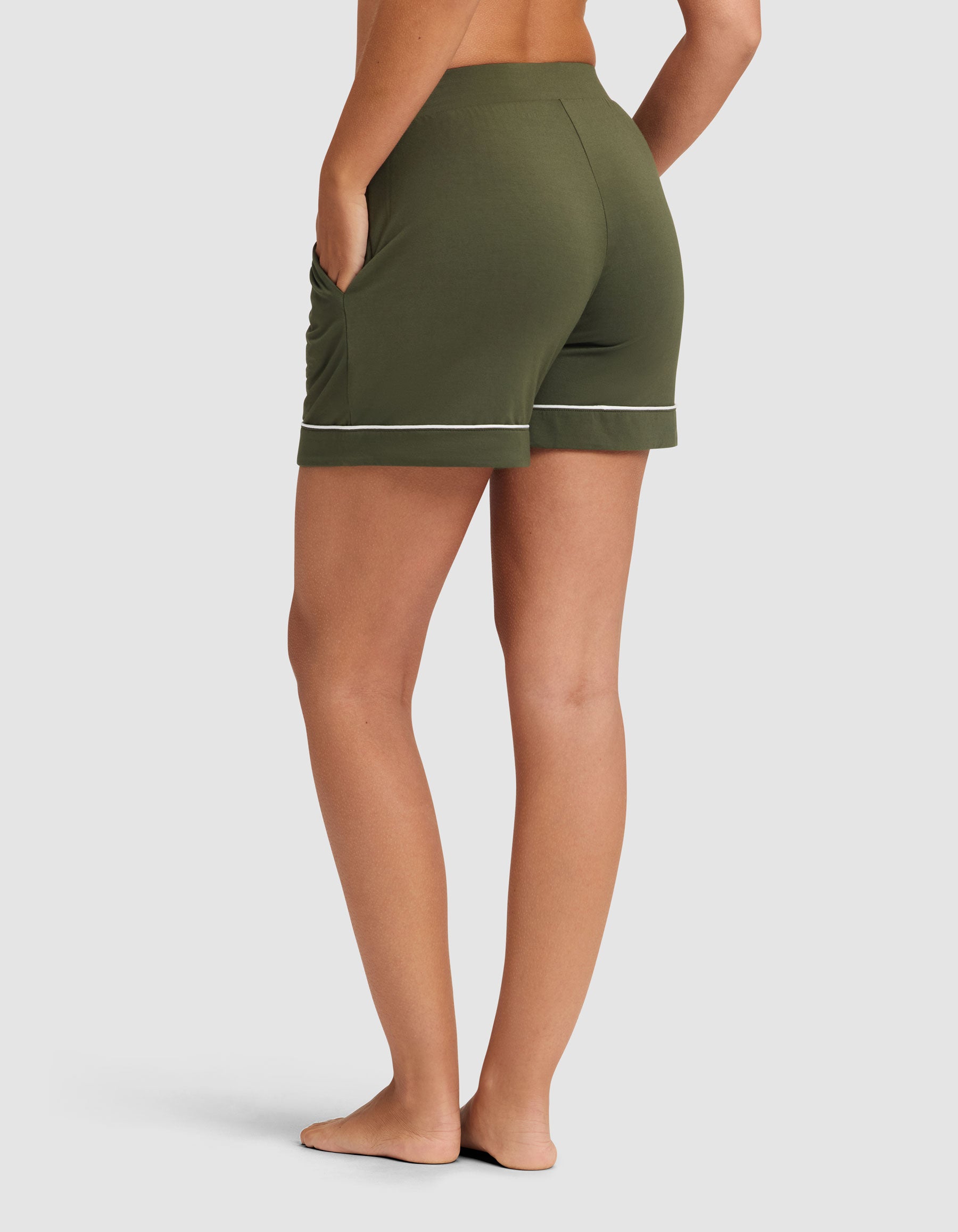 A woman models HIDE's Women's Bamboo Stretch-Knit Pajama Short in olive green with white piping, standing barefoot on a white background, viewed from the back with one hand in a pocket. 