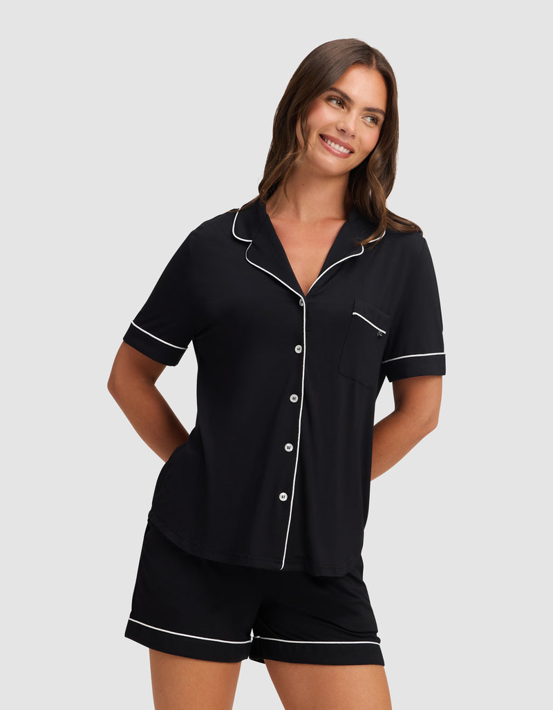 A woman smiles wearing HIDE's Women's Bamboo Stretch-Knit Short Sleeve Pajama Top in black with white piping, featuring a button-up front and chest pocket, paired with matching shorts against a plain light gray background.