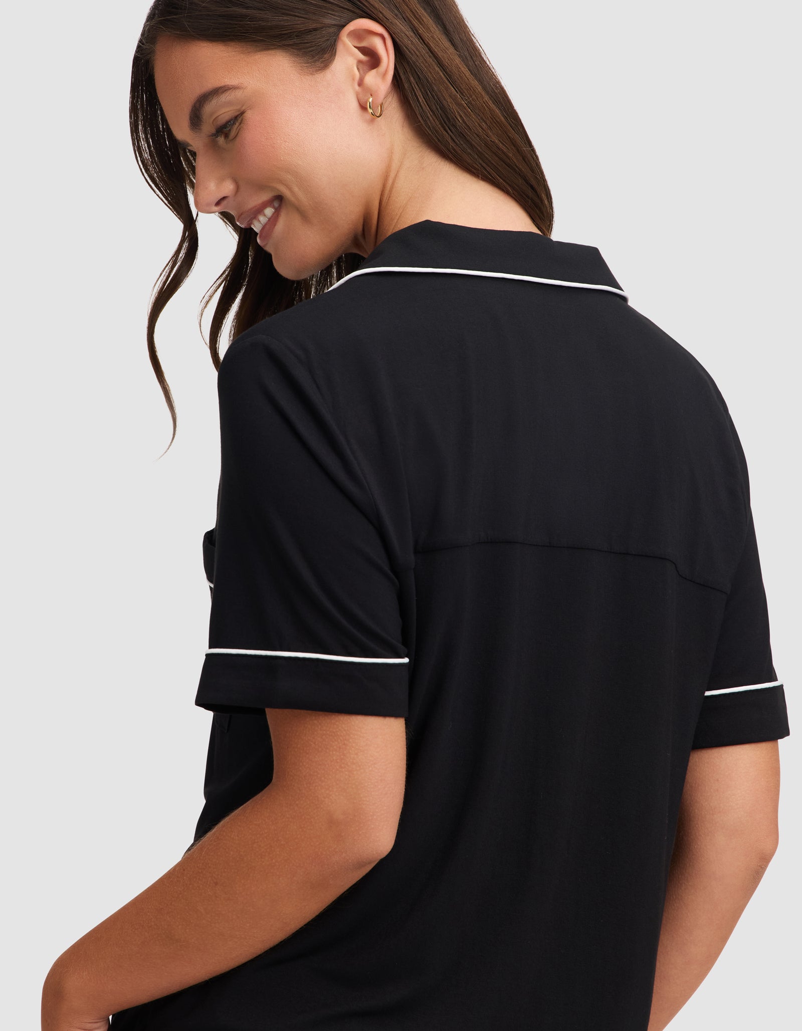 A woman with long brown hair smiles while looking down, wearing the HIDE Women's Bamboo Stretch-Knit Short Sleeve Pajama Top in black with white piping, shown from the back and side against a plain light gray background. 