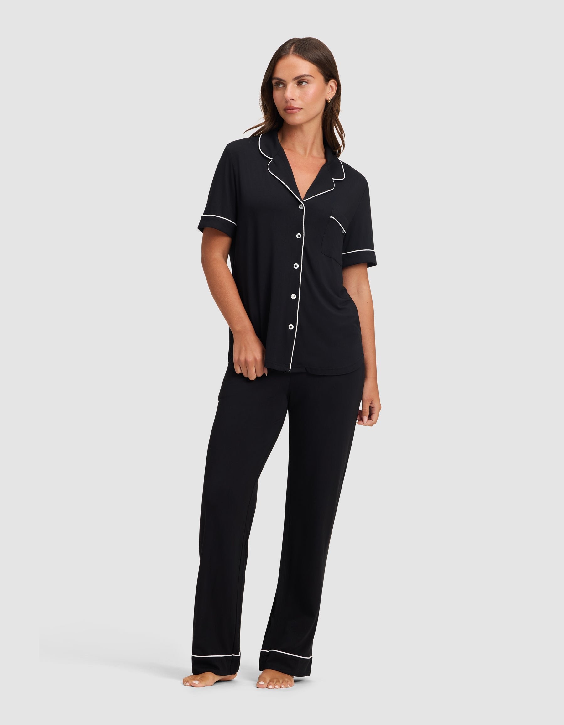 A woman in HIDE's Women's Bamboo Stretch-Knit Short Sleeve Pajama Top and matching black pants with white piping stands barefoot against a plain light background. 