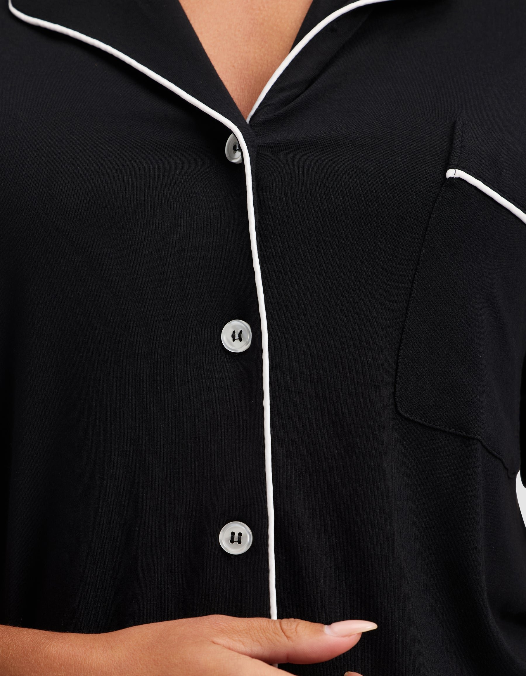A close-up shows a person wearing the HIDE Women's Bamboo Stretch-Knit Short Sleeve Pajama Top in black, featuring white piping on the collar, button placket, and pocket edge, with their hand visible near the bottom of the frame. 