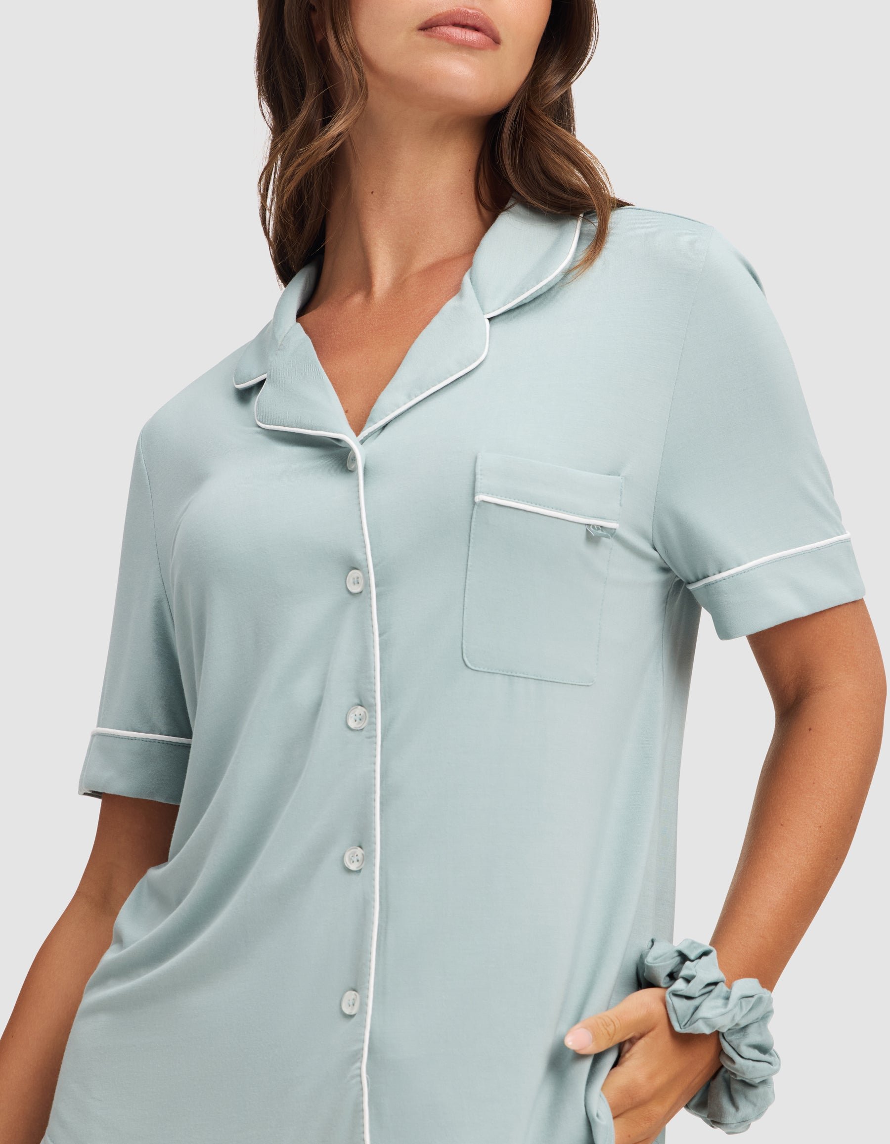A woman wears the HIDE Women's Bamboo Stretch-Knit Short Sleeve Pajama Top in light blue with white piping, buttons, and a chest pocket. She also has a matching scrunchie on her wrist. Her face is partially out of frame. 