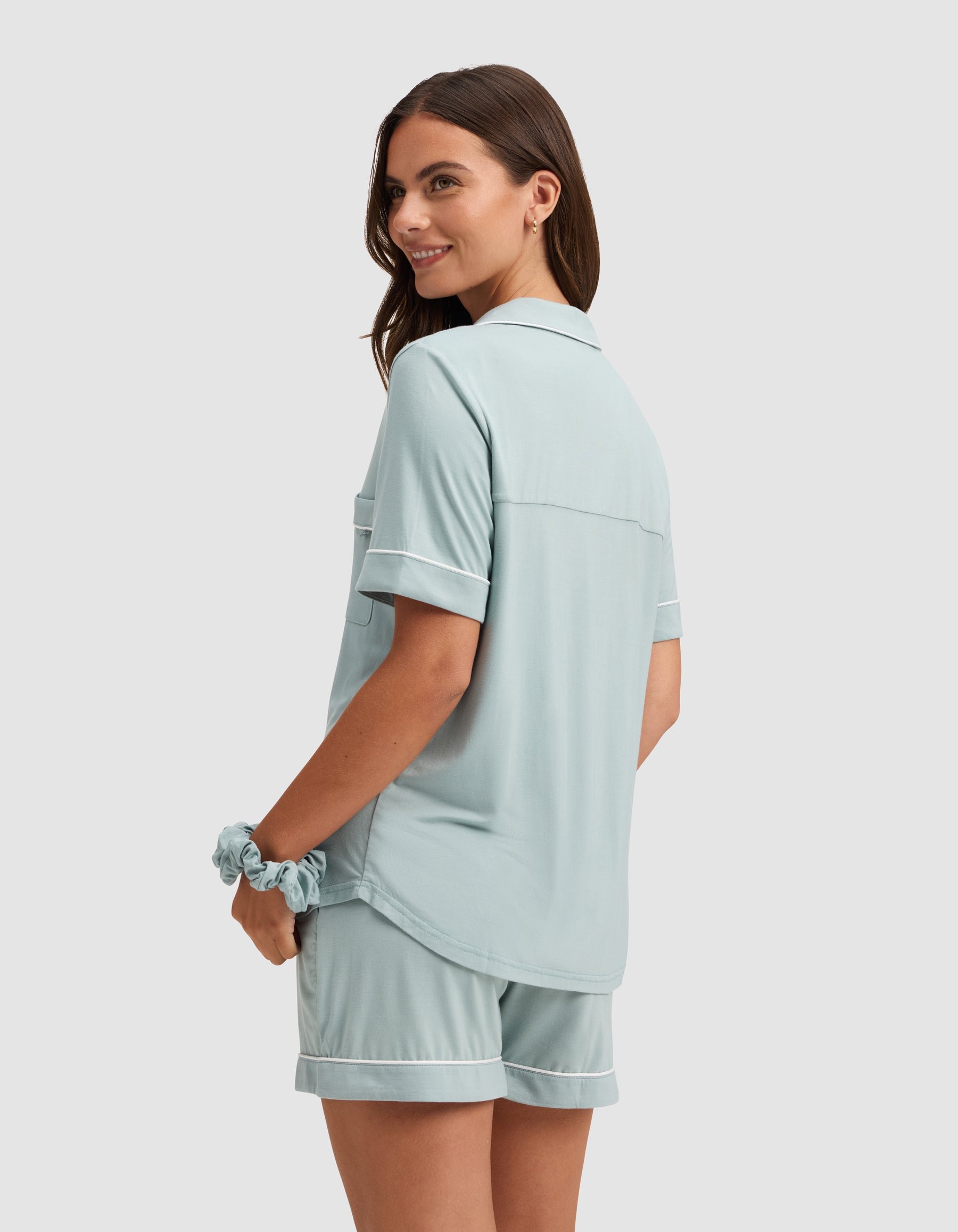 A woman smiles, standing sideways in a light blue HIDE Women's Bamboo Stretch-Knit Short Sleeve Pajama Top with matching shorts. She has long brown hair, a matching scrunchie on her wrist, and stands against a plain light background.  