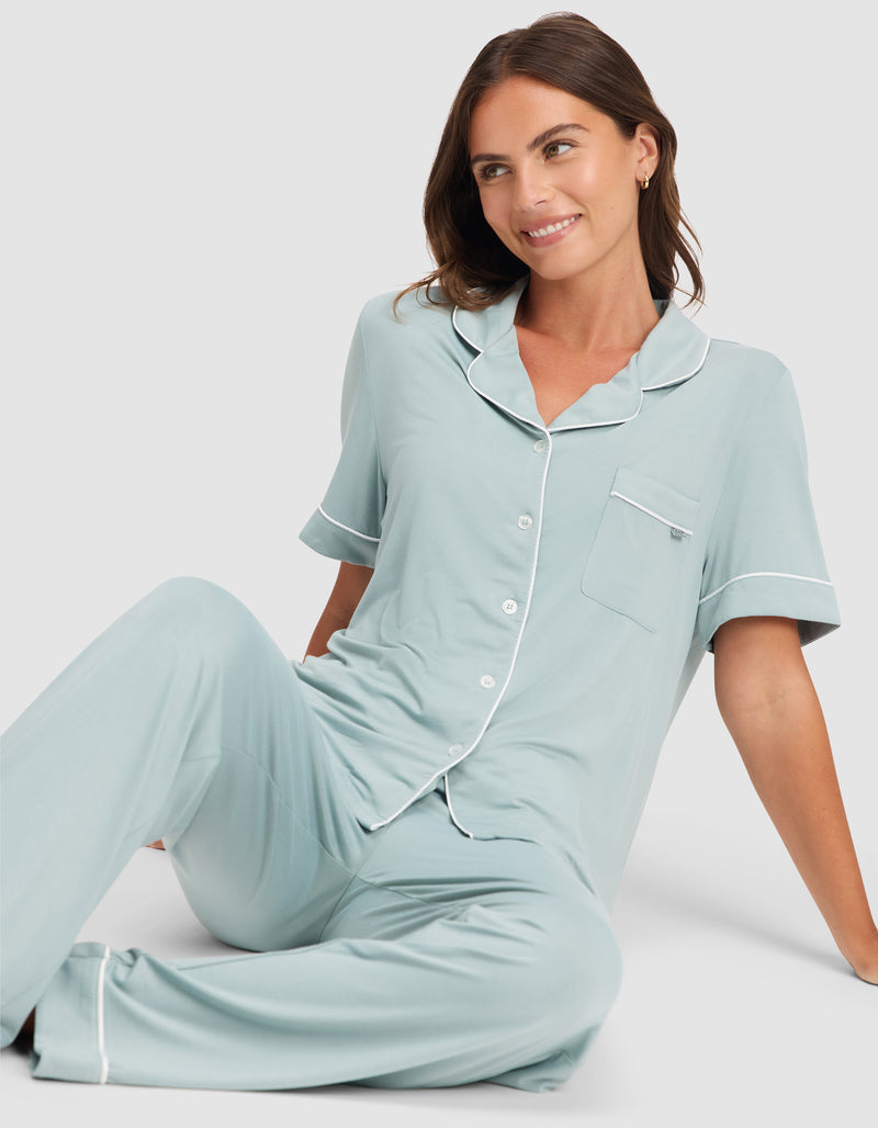 A woman with brown hair sits on the floor, smiling. She wears the HIDE Women's Bamboo Stretch-Knit Short Sleeve Pajama Top in light blue with white trim and matching long pants. The background is plain and light-colored.
