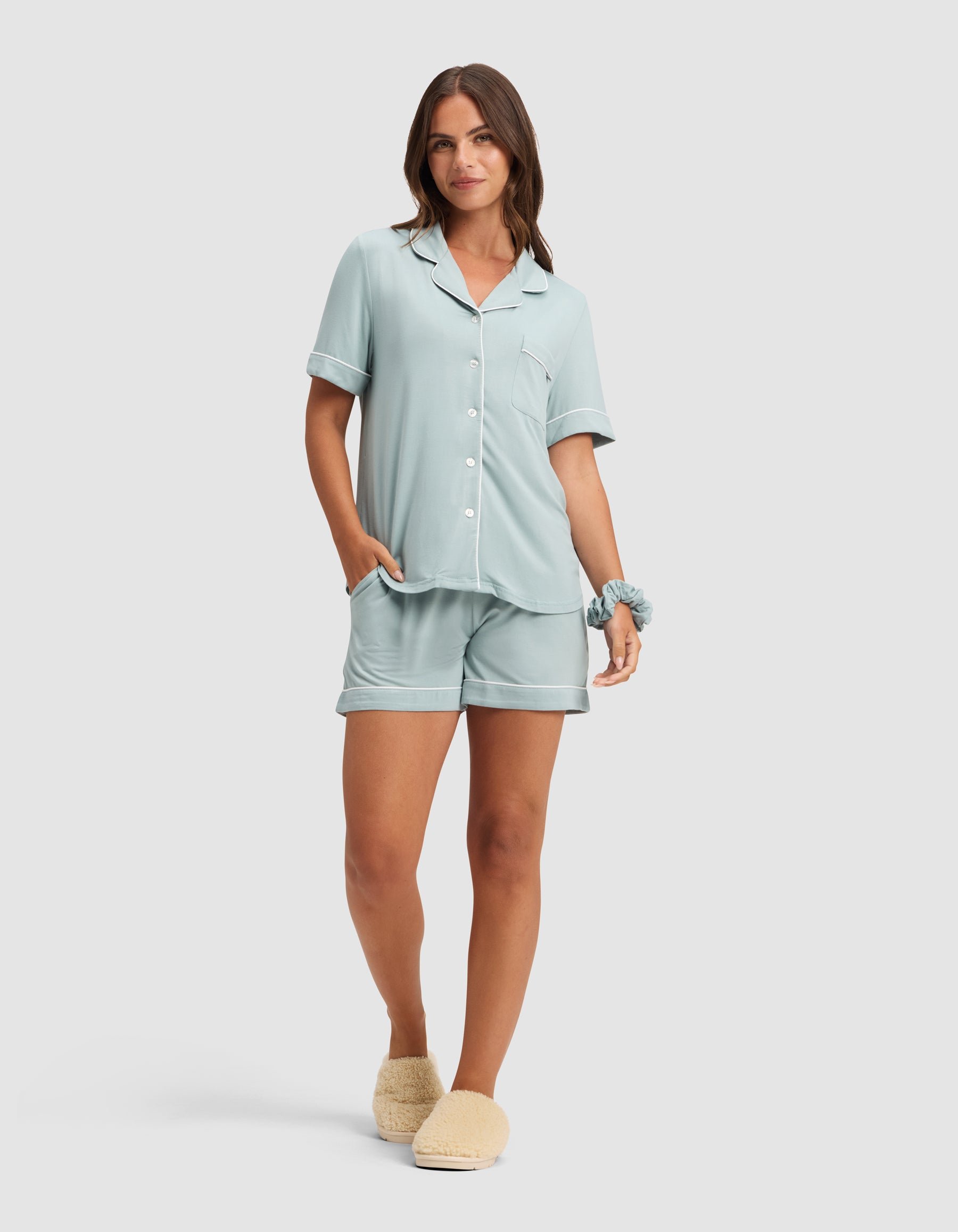 A woman smiles with her hands in her pockets, wearing the HIDE Women's Bamboo Stretch-Knit Short Sleeve Pajama Top in light blue, matching shorts, a scrunchie on her wrist, and beige slippers. 