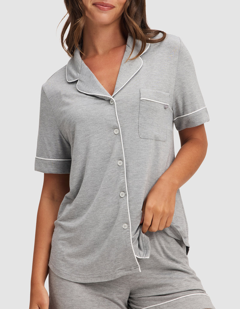 A woman wears the HIDE Women's Bamboo Stretch-Knit Short Sleeve Pajama Top in light gray with matching shorts, featuring white piping, a chest pocket, and button-down front, against a plain background.