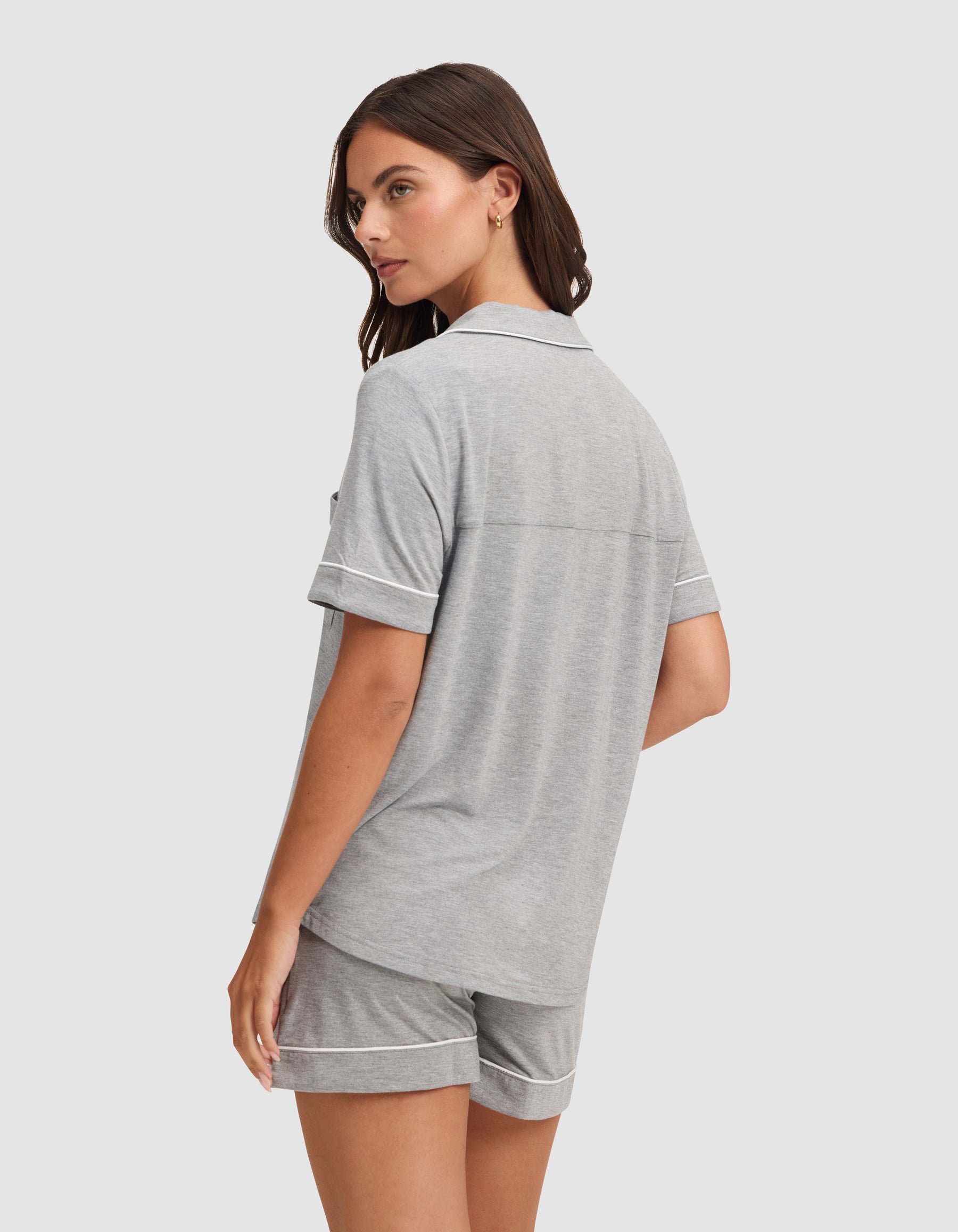 A woman with brown hair models the HIDE Women's Bamboo Stretch-Knit Short Sleeve Pajama Top in light gray with white trim, shown from the back as she looks over her shoulder against a plain light background. 