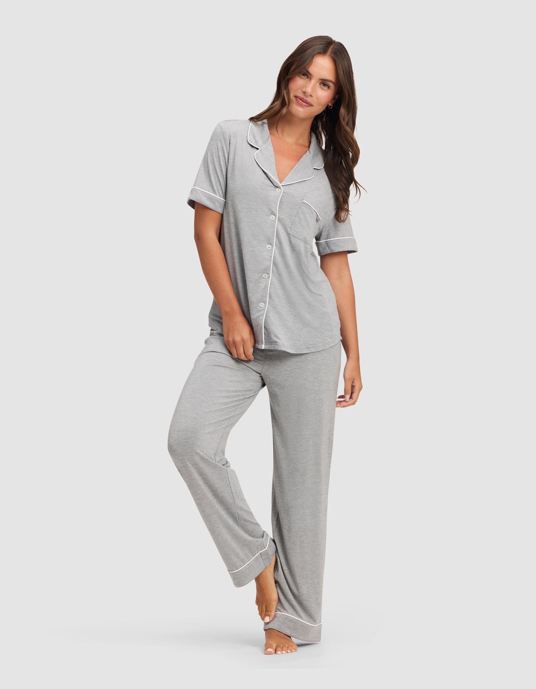 A woman with long brown hair stands barefoot against a plain background, wearing the HIDE Women's Bamboo Stretch-Knit Short Sleeve Pajama Top in light gray with white trim and button-up front, paired with matching long pants. 