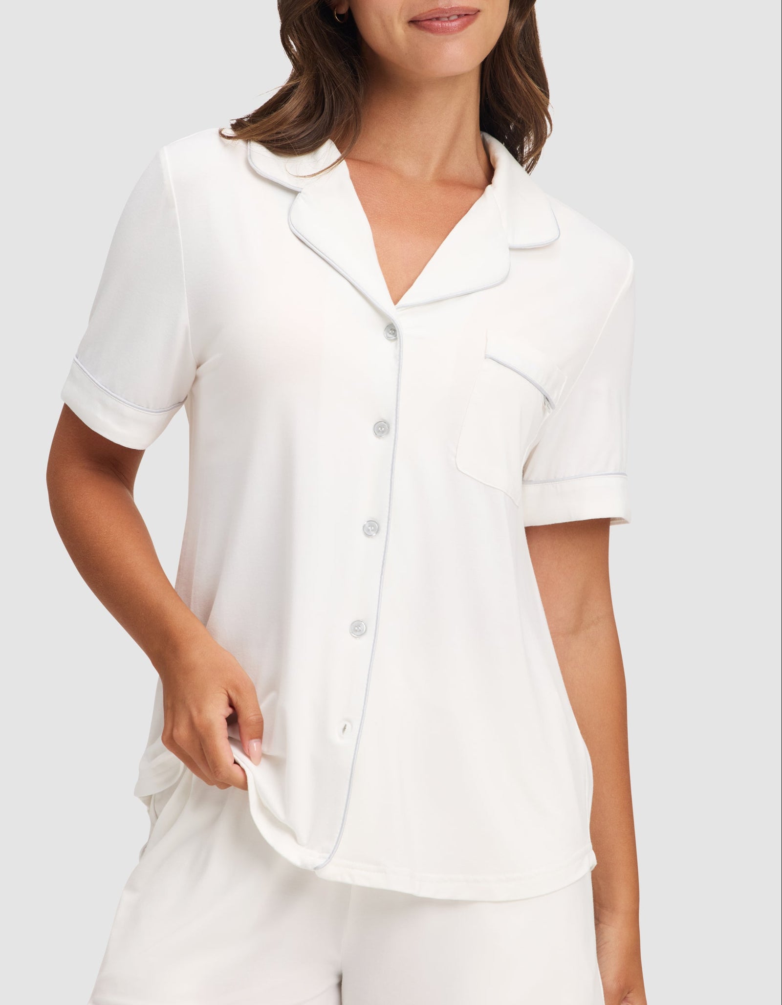 A woman wears the HIDE Women's Bamboo Stretch-Knit Short Sleeve Pajama Top with buttons and a chest pocket, paired with matching white shorts, standing against a plain light background. 
