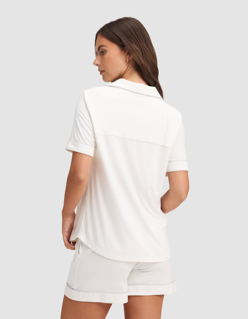 A woman with long brown hair is standing slightly turned away, wearing the HIDE Women's Bamboo Stretch-Knit Short Sleeve Pajama Top in white, set against a plain light background.