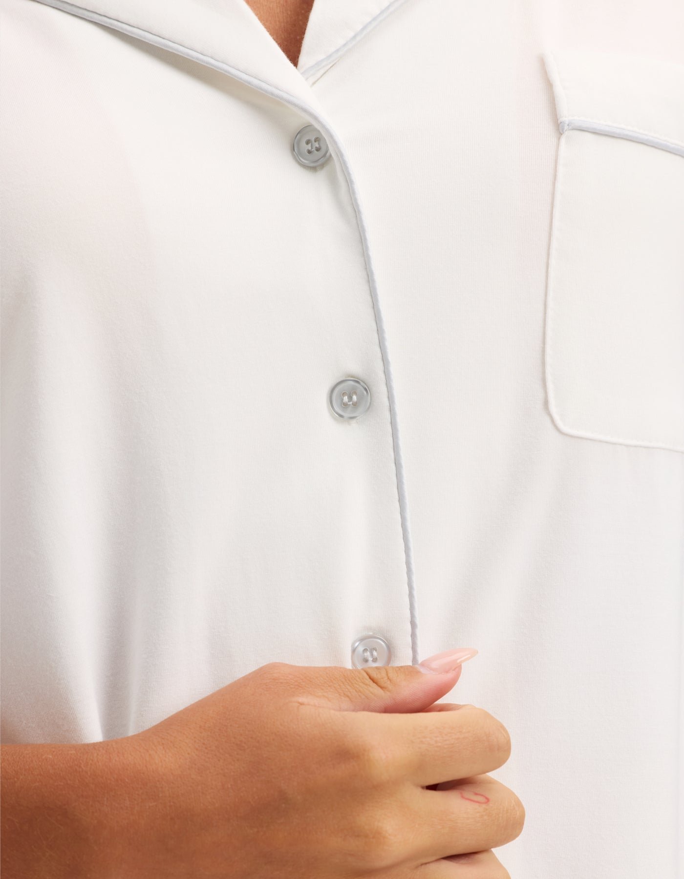 A close-up of a person wearing HIDE's Women's Bamboo Stretch-Knit Short Sleeve Pajama Top, holding the fabric near the buttons with their left hand, showing pale pink manicured nails. 