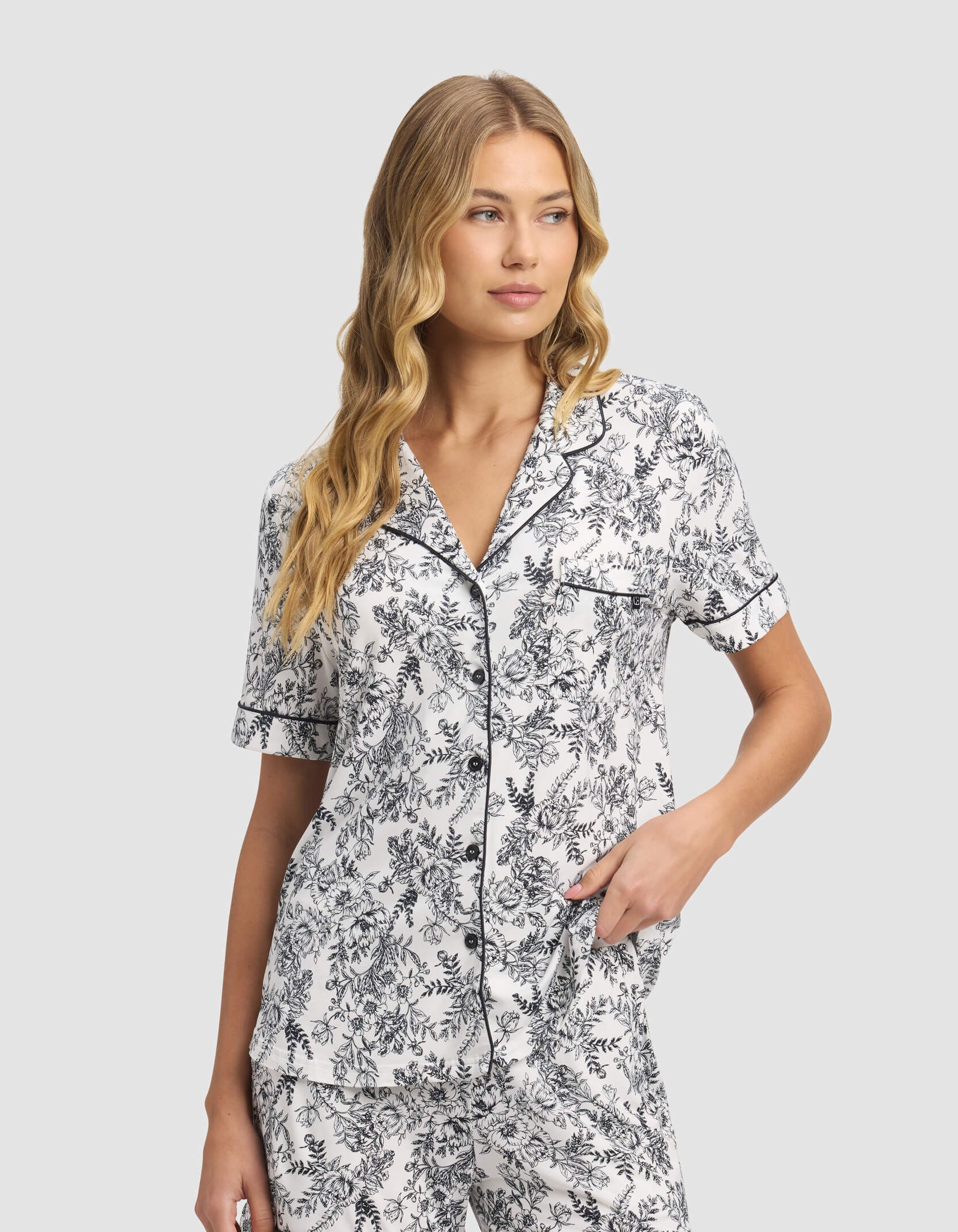 A woman with long, wavy blonde hair models the Cozy Earth Women's Bamboo Stretch-Knit Short Sleeve & Pant Pajama Set in a black and white floral print, standing and looking to her left with a neutral expression.|Color:Jet Black Toile