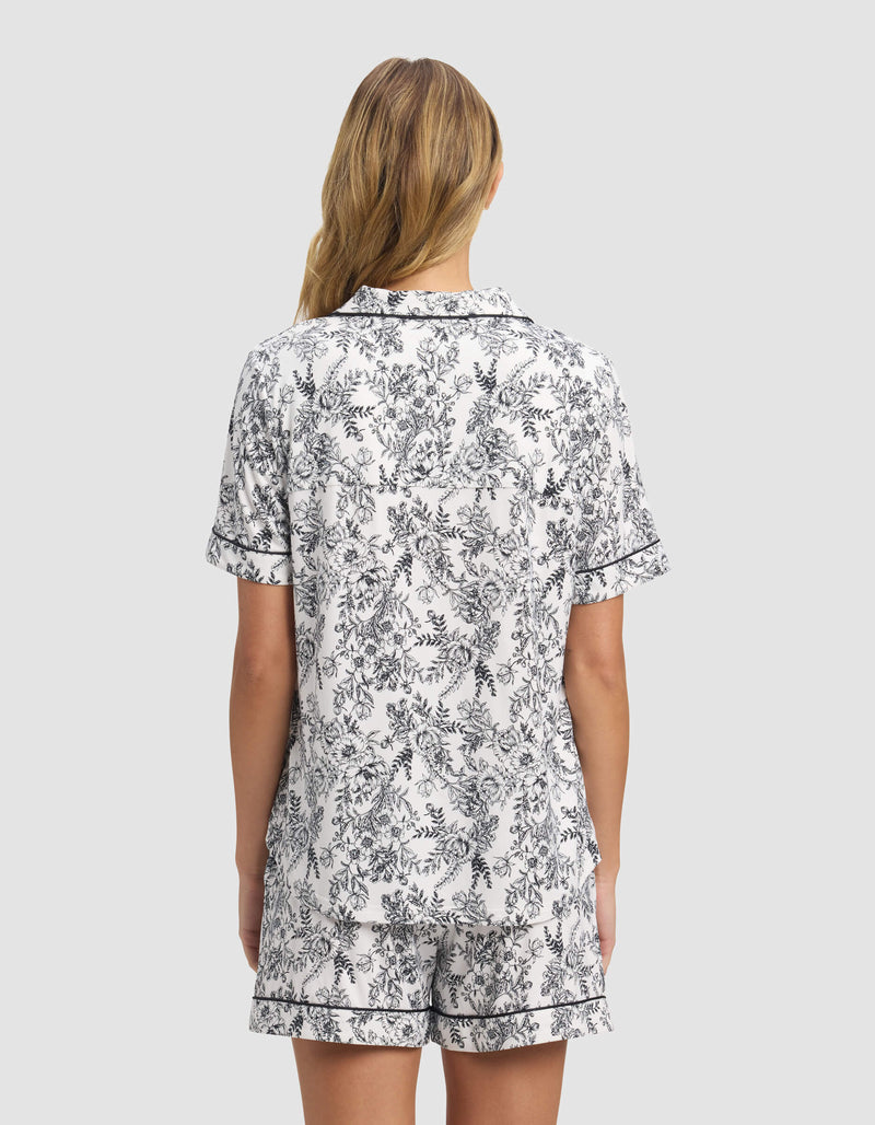 A woman with long, blonde hair is seen from behind wearing Cozy Earth's Women's Bamboo Stretch-Knit Short Sleeve Pajama Top and matching shorts with a black and white floral print, against a plain light gray background.