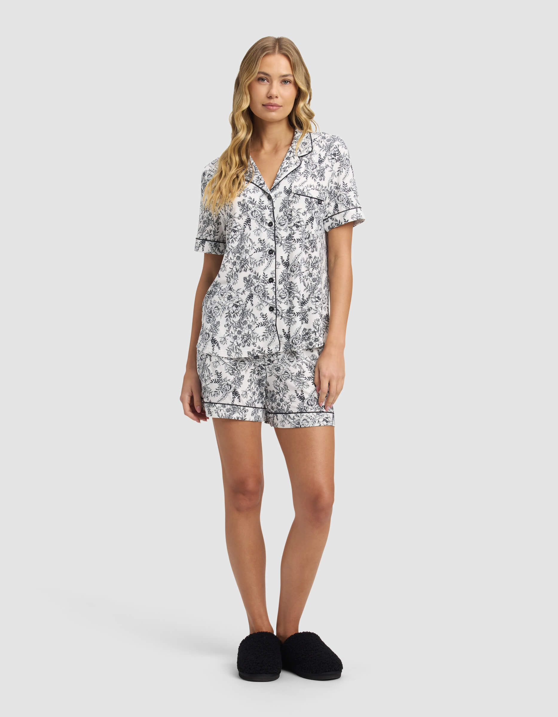 A woman with long blonde hair wears the Cozy Earth Women's Bamboo Stretch-Knit Short Sleeve Pajama Top, featuring a black and white floral pattern, paired with black slippers. She stands against a plain light gray background.