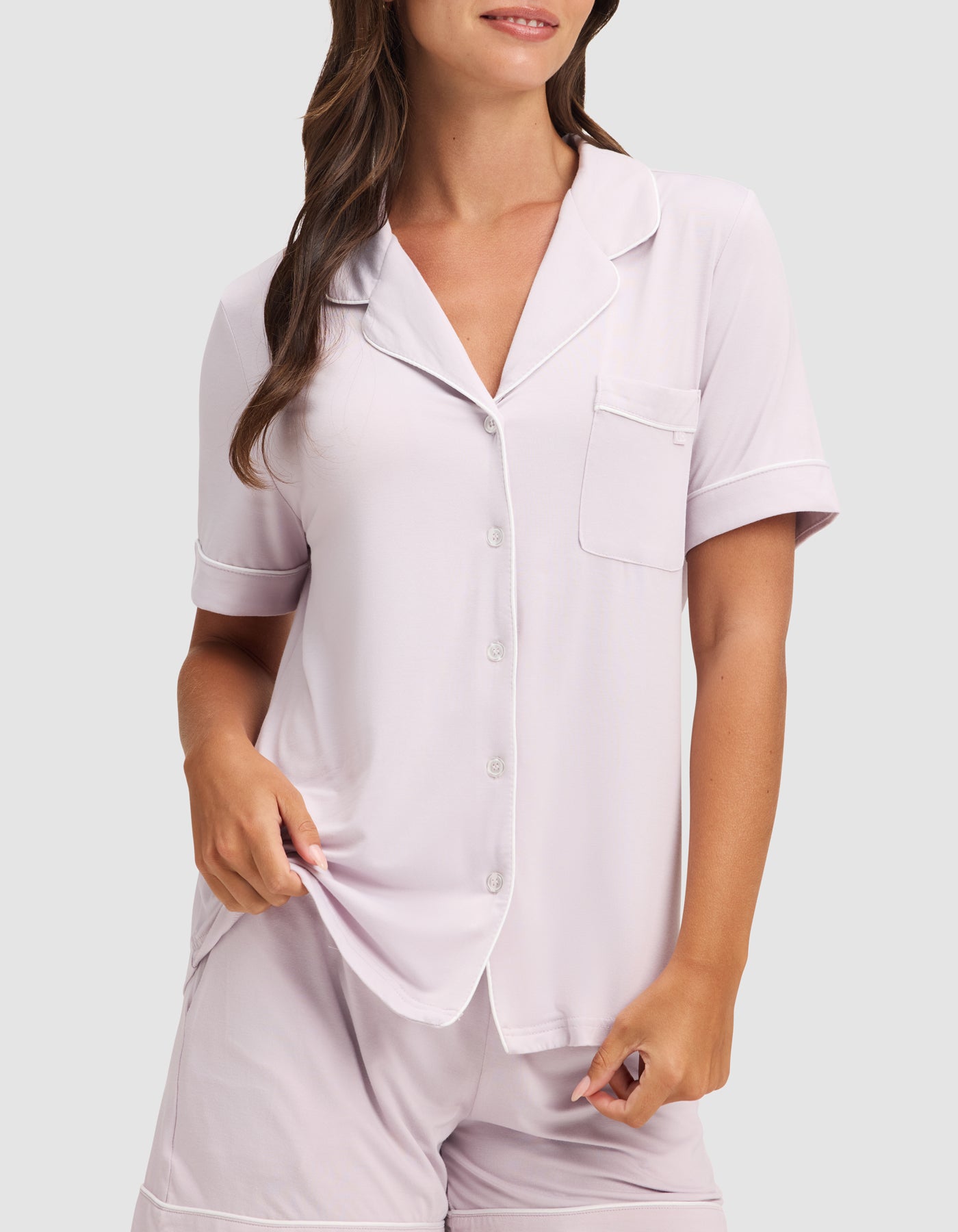 A woman stands against a plain light gray background wearing the HIDE Women's Bamboo Stretch-Knit Short Sleeve Pajama Top in light pink with a breast pocket and matching shorts. 