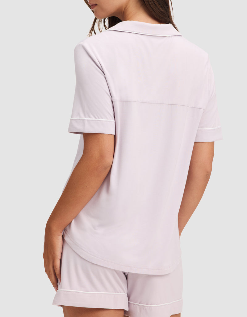 A woman is shown from the back wearing HIDE's Women's Bamboo Stretch-Knit Short Sleeve Pajama Top in light pink with matching shorts, standing against a plain light gray background.