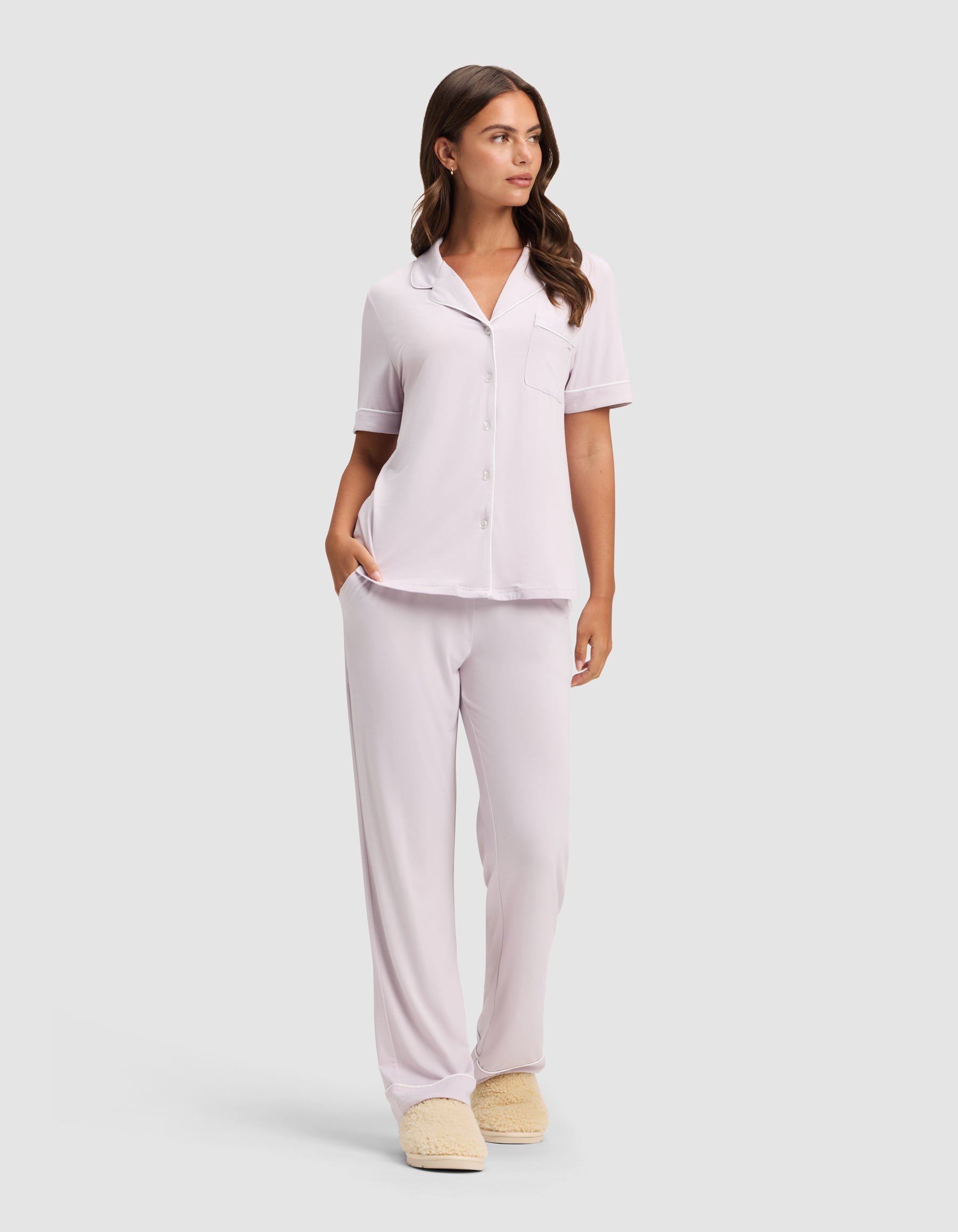 A woman wears the HIDE Women's Bamboo Stretch-Knit Short Sleeve Pajama Top in light pink with matching pants and beige slippers, standing against a plain light gray background with one hand in her pocket and looking to the side. 